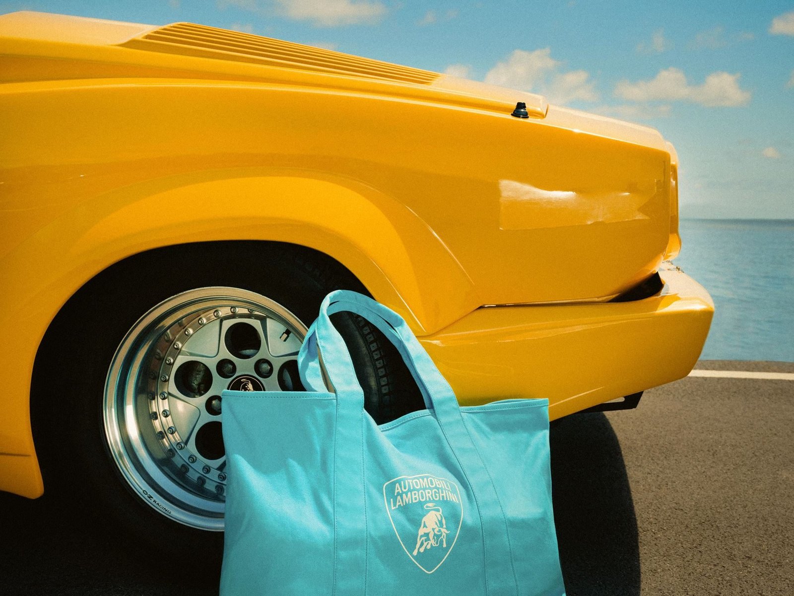 Yellow Lamborghini Countach parked by the sea with a light blue Automobili Lamborghini tote bag in the foreground