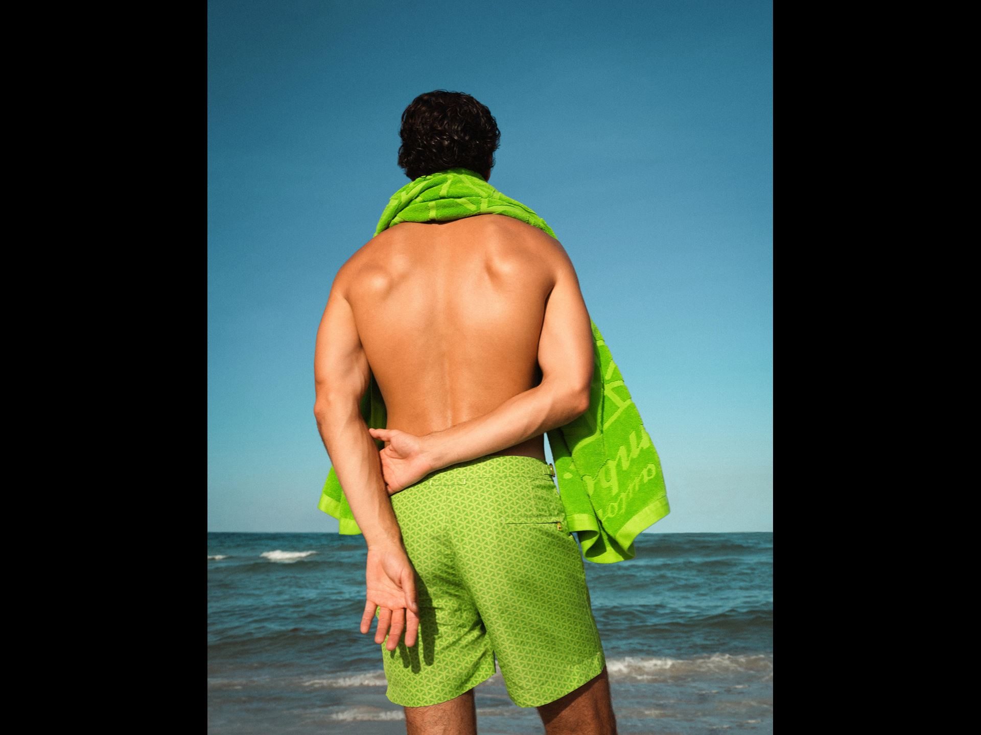 Lamborghini'S Orlebar Brown Beach Collection Turns Countach Lines And Ad Personam Colors Into Luxury Swimwear 3 Man wearing green lamborghini-branded towel and green patterned swim shorts looking out at the ocean