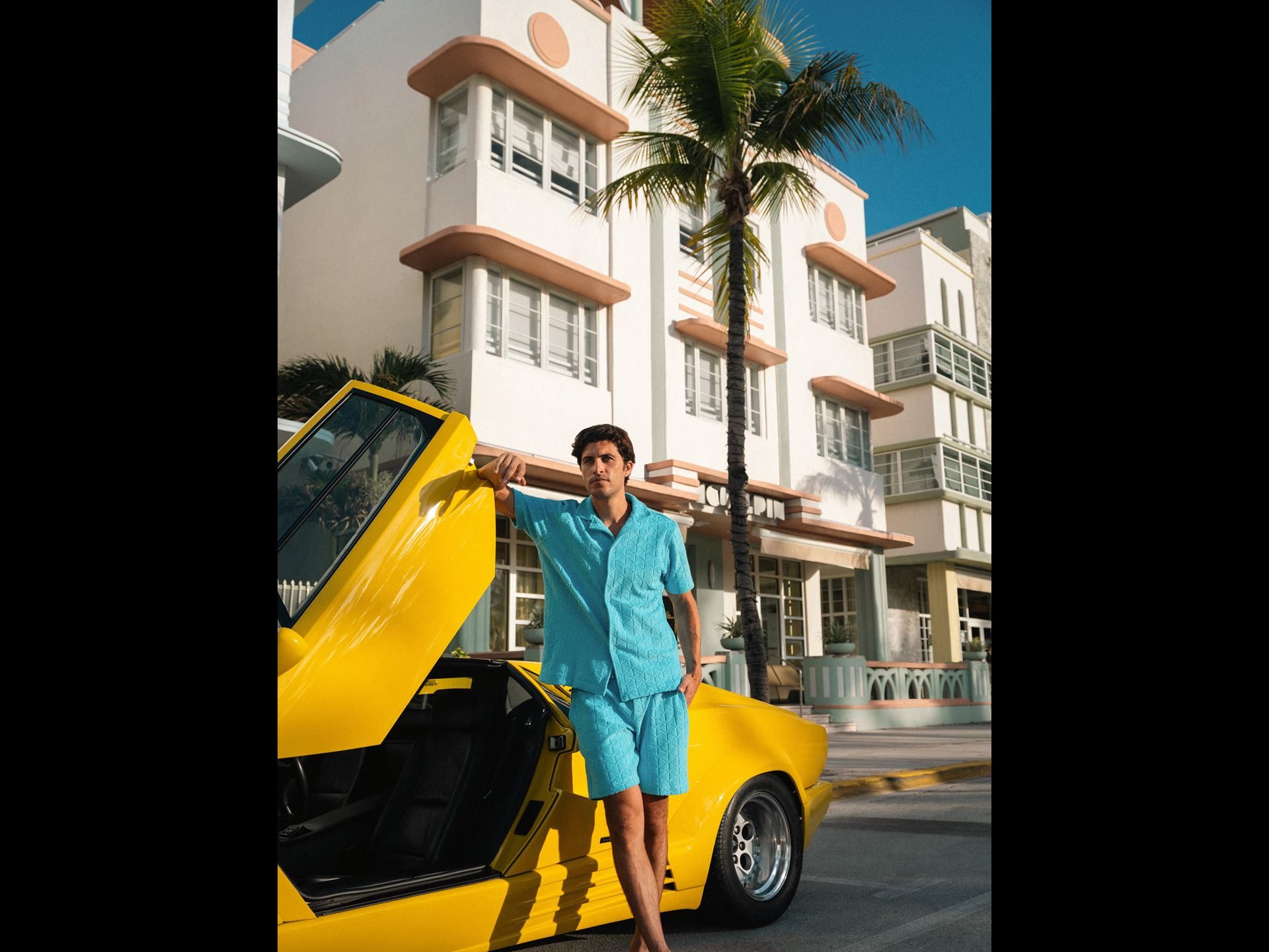 Lamborghini'S Orlebar Brown Beach Collection Turns Countach Lines And Ad Personam Colors Into Luxury Swimwear 5 Man in light blue patterned orlebar brown shirt and shorts leaning against the open scissor door of a yellow lamborghini countach 25th anniversary in front of an art deco building