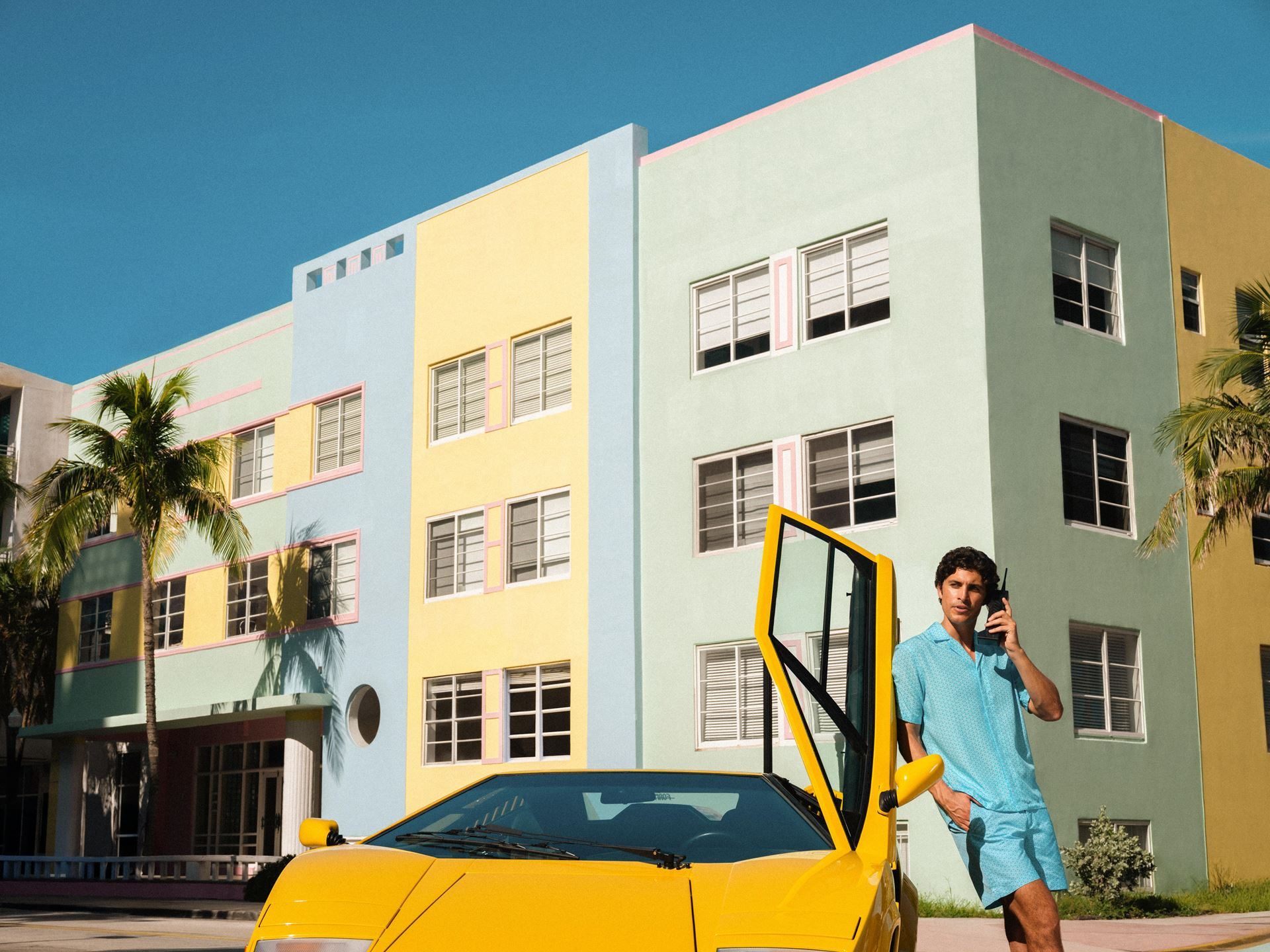 Lamborghini'S Orlebar Brown Beach Collection Turns Countach Lines And Ad Personam Colors Into Luxury Swimwear 7 Man in light blue patterned orlebar brown shirt holding a vintage mobile phone, leaning against the open scissor door of a yellow lamborghini countach 25th anniversary in front of colorful art deco buildings