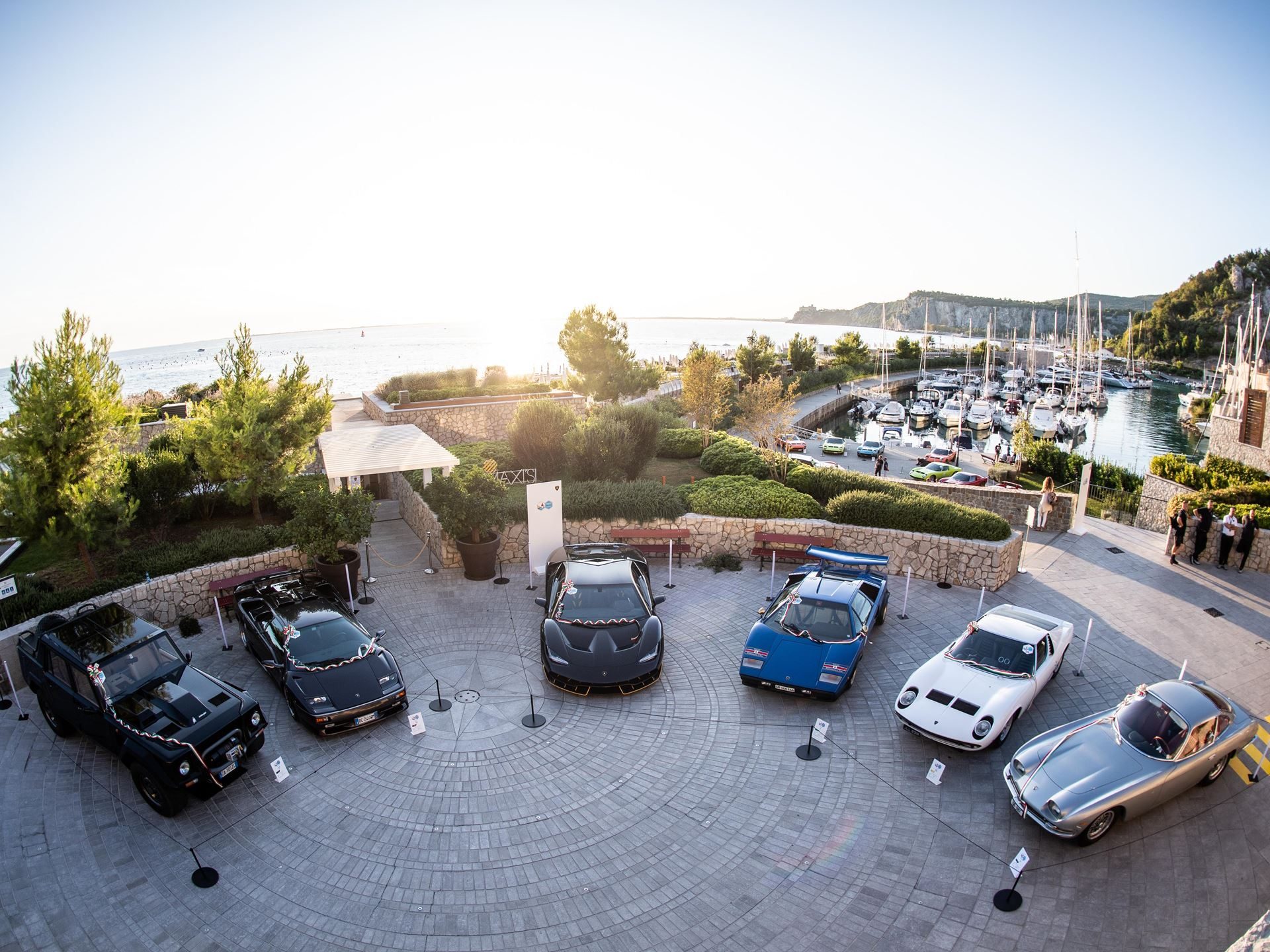 Ten Years Of Polo Storico: How Lamborghini Built The Only Heritage Program That Rebuilds Cars From The Dead 13 Circular display of classic lamborghini cars including miura, countach, and lm002 at a coastal resort at sunset