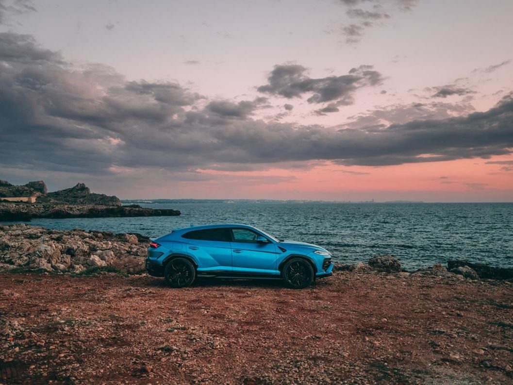A blue lamborghini urus se parked on a rocky coastline overlooking the sea at sunset
