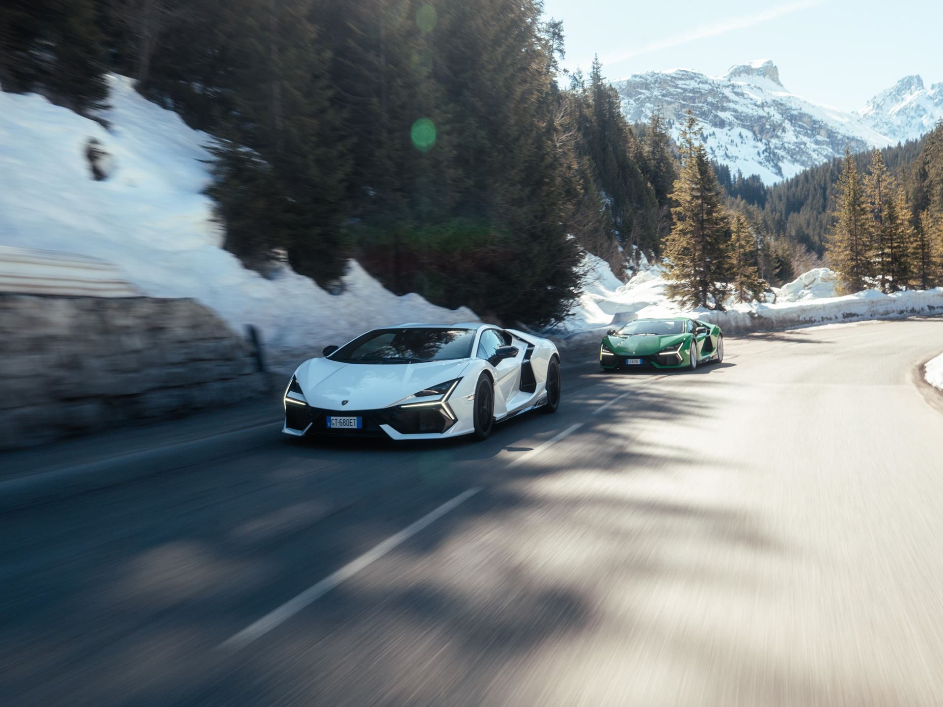 Lamborghini Took Its Hybrid Lineup To The French Alps, And The Convoy Included A Surprise Guest 11 Two lamborghini revuelto supercars, one white and one green, driving on a winding snowy mountain road with trees and sunlight