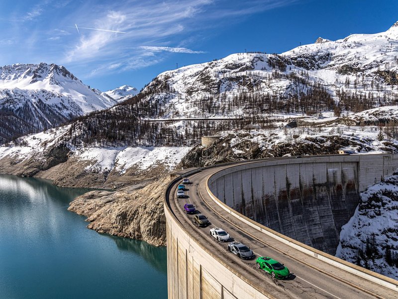 Convoy of Lamborghini Revuelto and Urus models driving on a curved dam road with snowy Alps and a lake in the background