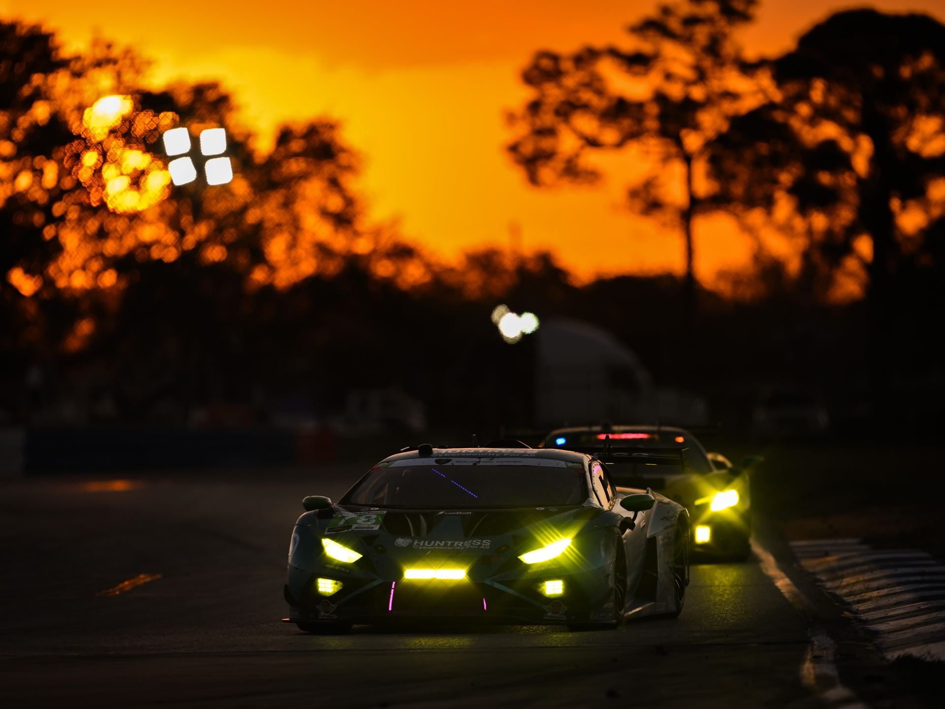 Kvyat Rejoins Grosjean And Mortara As The Lamborghini Sc63 Heads To Its Last Imsa Gtp Start 7 Two lamborghini huracán gt3 evo2 race cars with bright yellow headlights racing at dusk against a dramatic orange sky