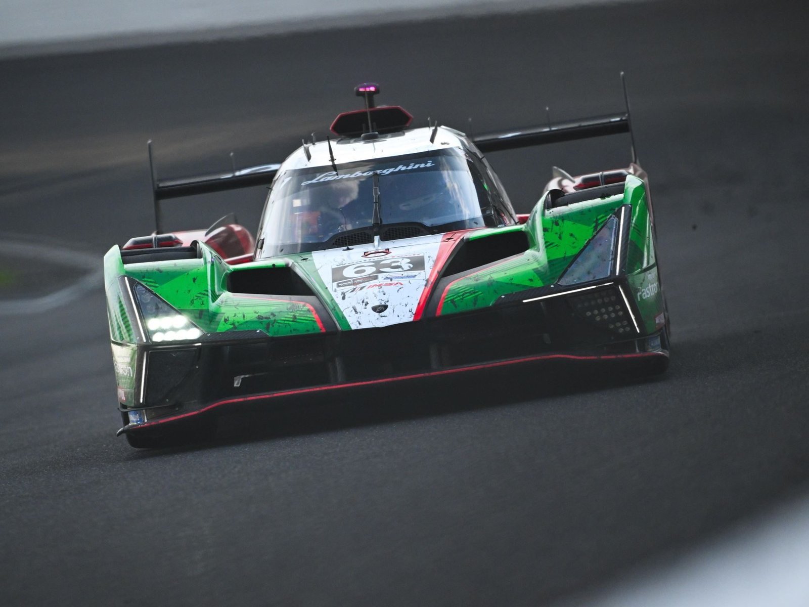 Lamborghini SC63 LMDh race car in green, white, and red livery racing on a dark circuit with LED headlights illuminated