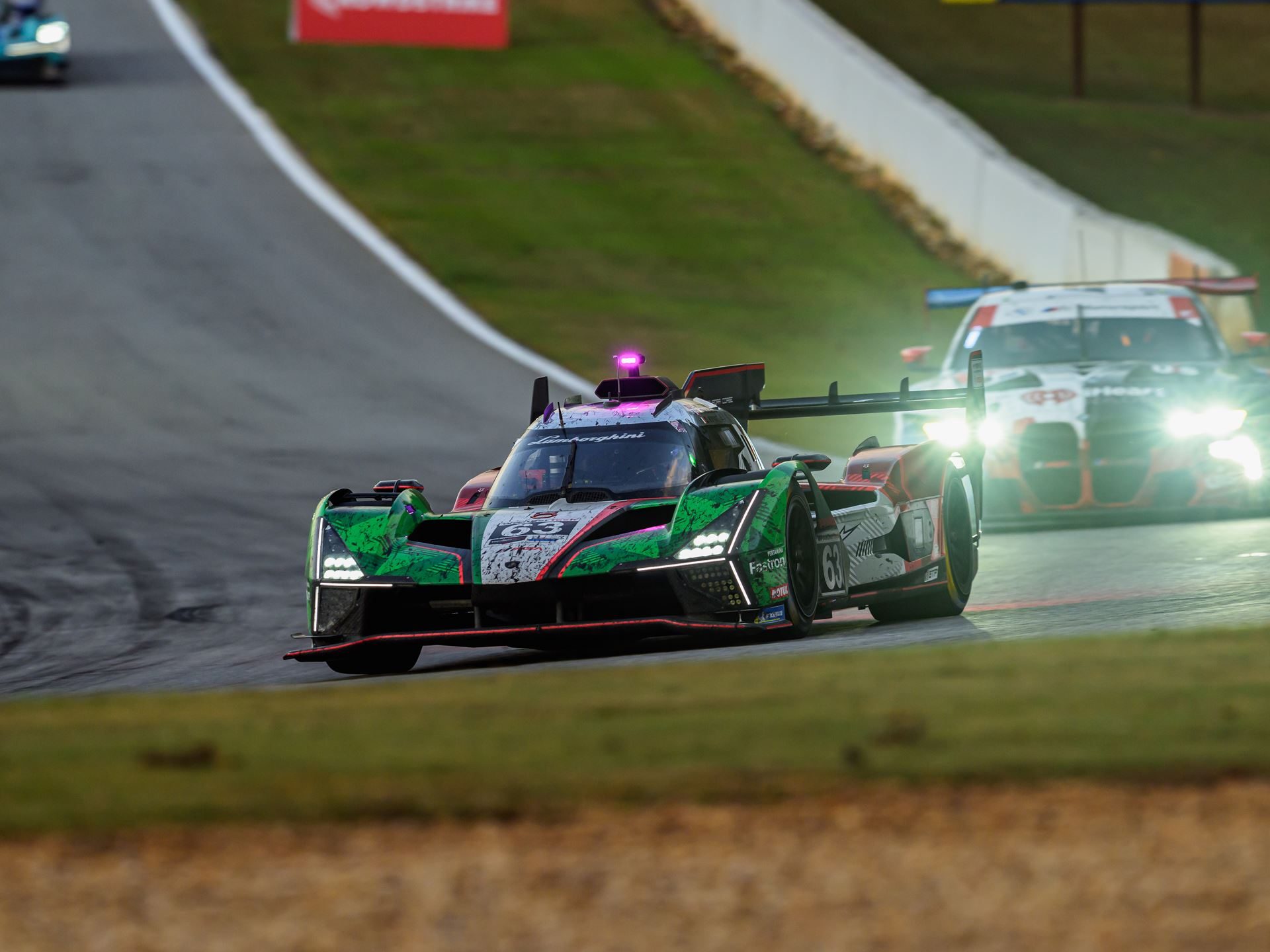The lamborghini sc63 leads a competitor on track at road atlanta as dusk approaches with headlights illuminated