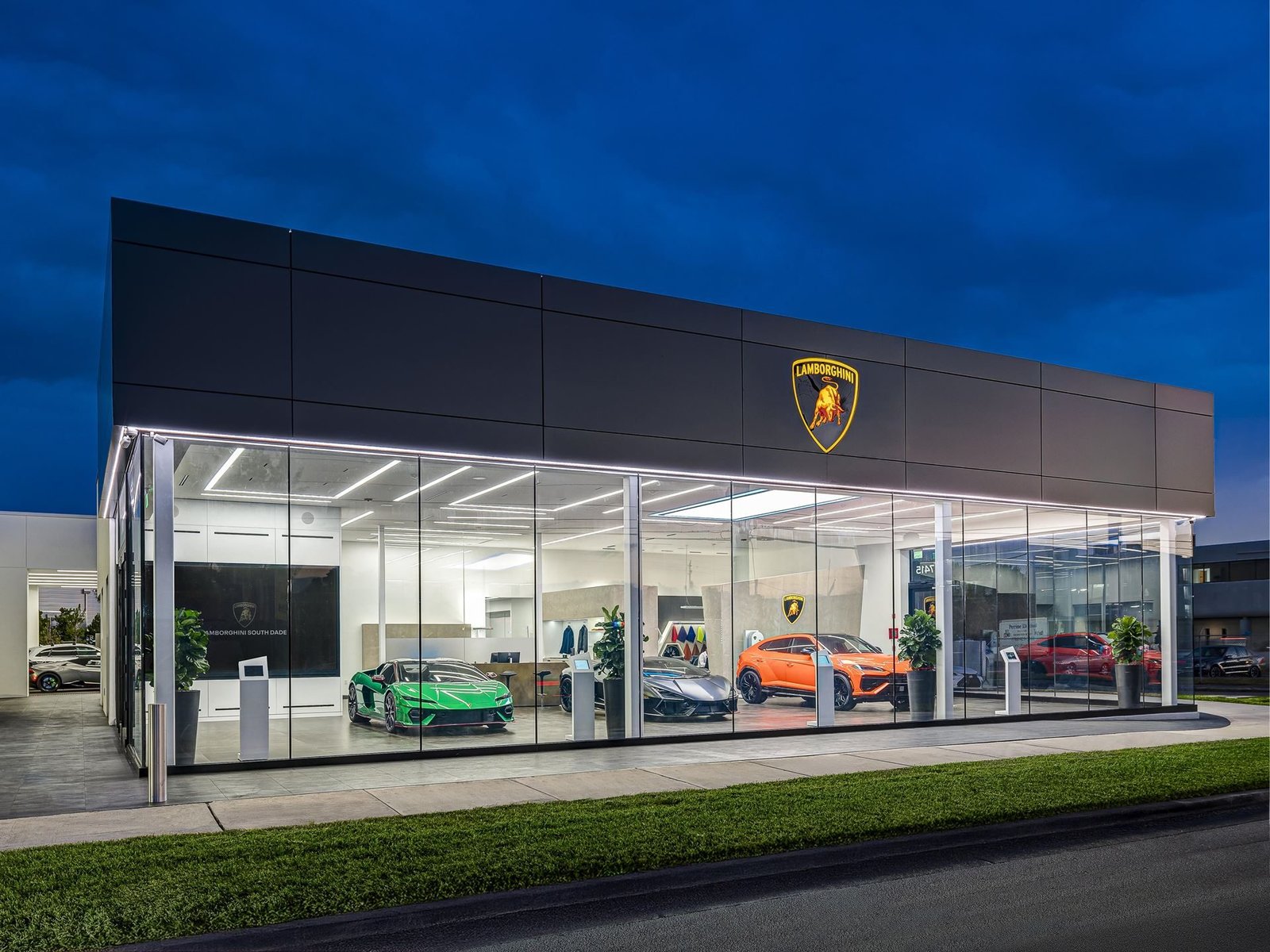 Lamborghini South Dade showroom illuminated at dusk with a green Huracán, dark Revuelto, and orange Urus visible through glass windows