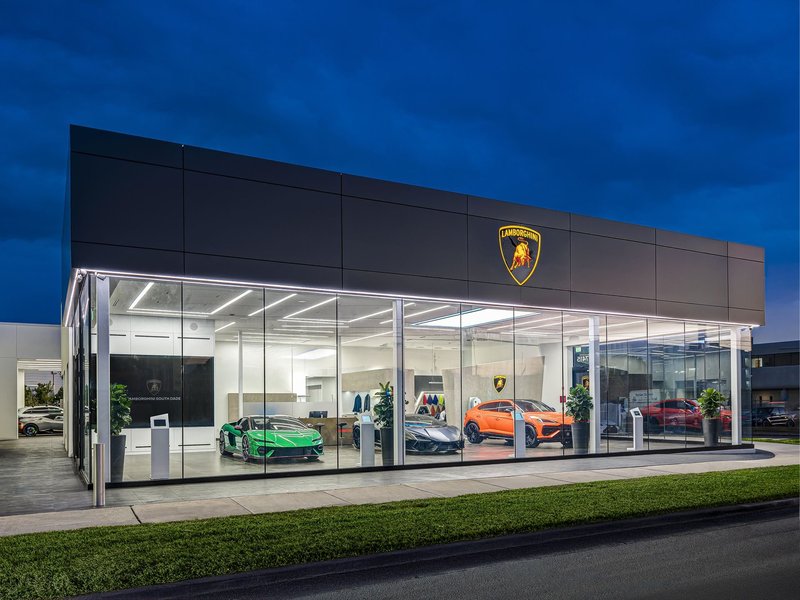 Lamborghini South Dade showroom illuminated at dusk with a green Huracán, dark Revuelto, and orange Urus visible through glass windows