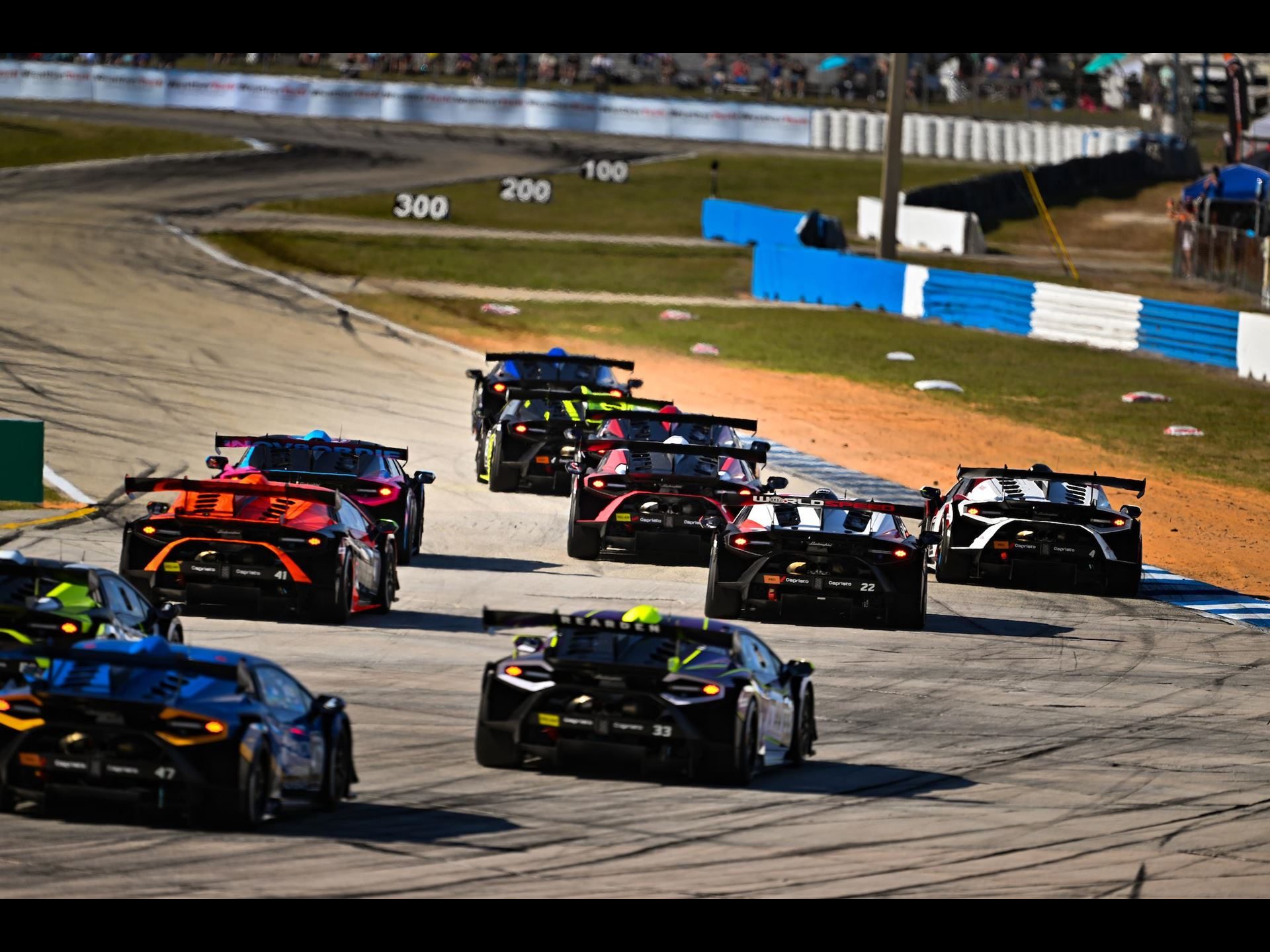41 Huracáns Head To Laguna Seca As Wayne Taylor Racing Carries Five-Win Sebring Momentum 7 A line of lamborghini huracán super trofeo evo2 race cars in various liveries navigating a turn, viewed from behind and above with spectators in the background