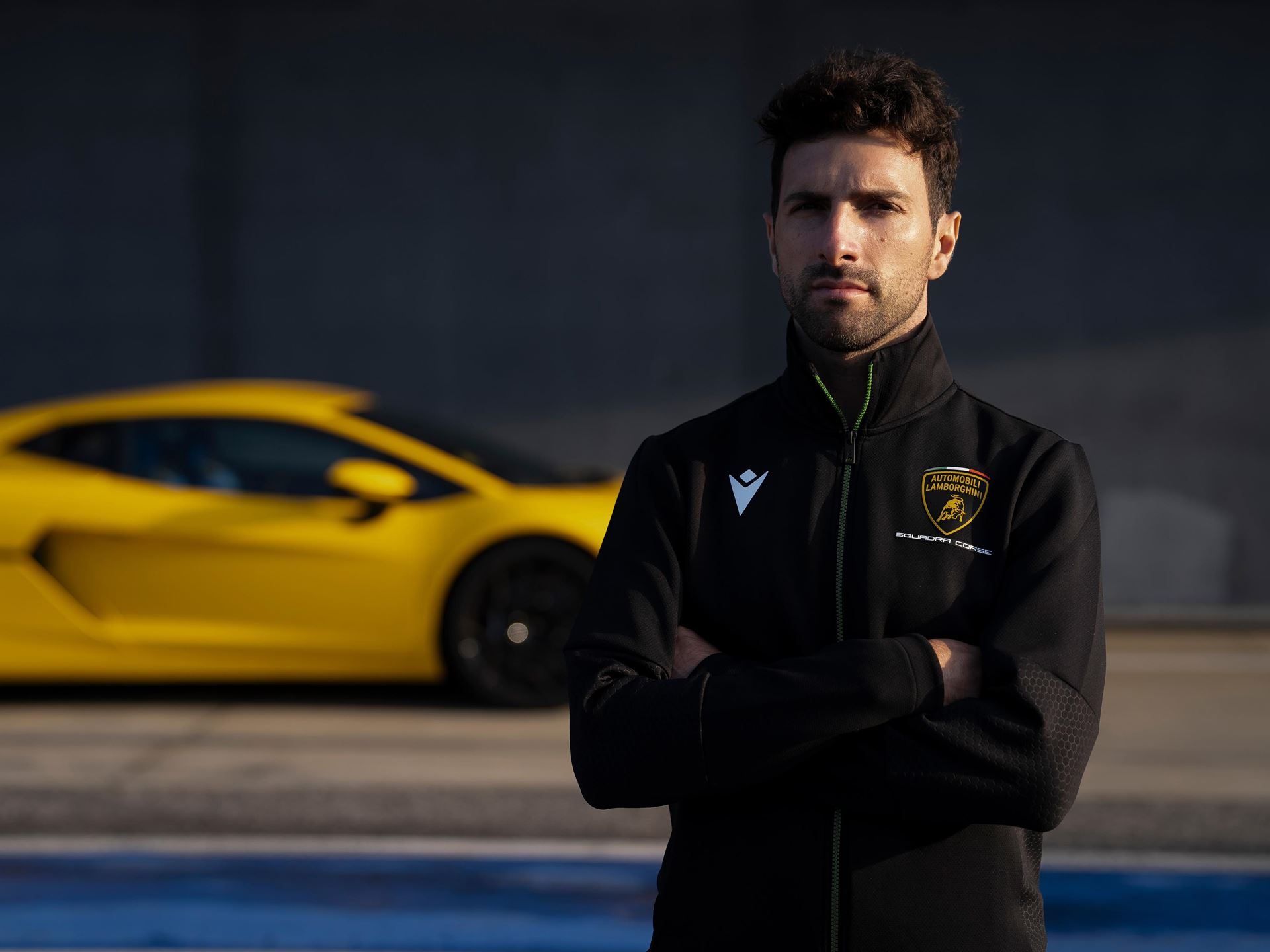Andrea caldarelli in lamborghini squadra corse jacket with a blurred yellow temerario on track behind him