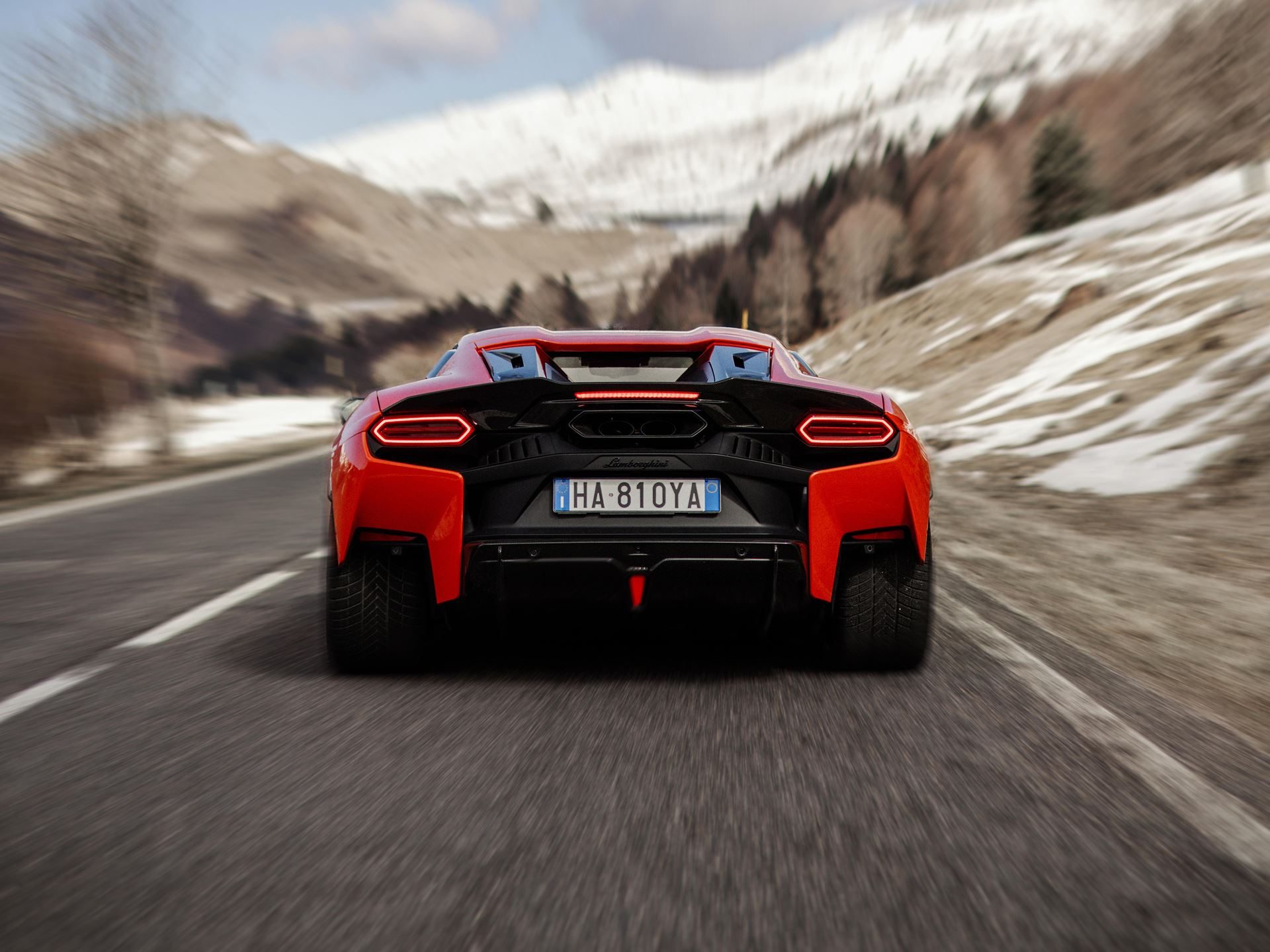 Rear view of an orange lamborghini temerario driving on a winding mountain road with motion blur, showing distinctive rear lights and central exhaust