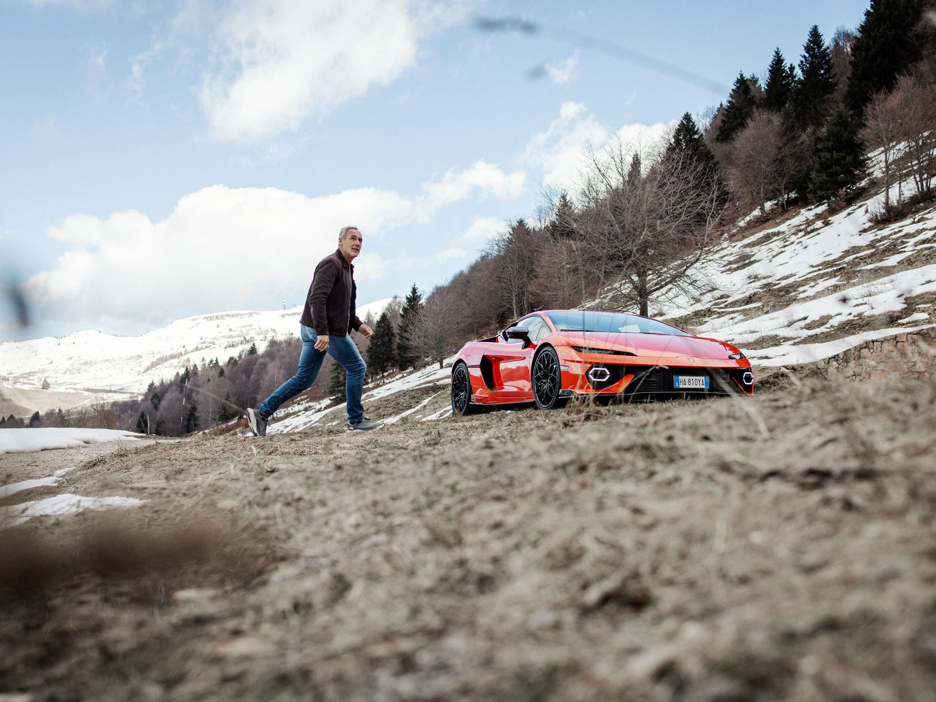 Mike horn walking towards an orange lamborghini temerario on a dirt path in a snowy mountainous landscape