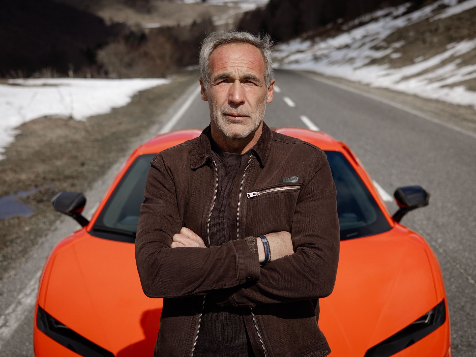 Mike horn standing with crossed arms in front of an orange lamborghini temerario on a snowy mountain road