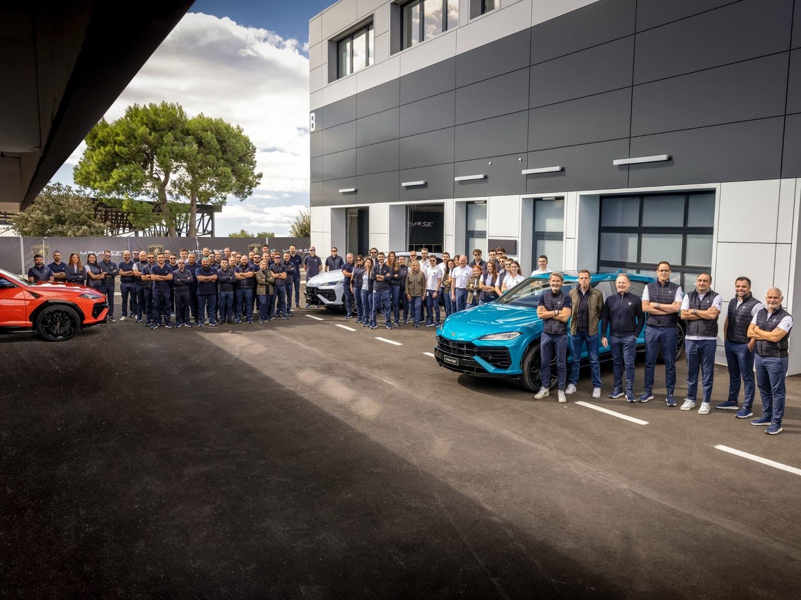 Large group of Lamborghini employees standing in front of the Sant'Agata Bolognese facility with three Lamborghini Urus models in orange, blue, and white