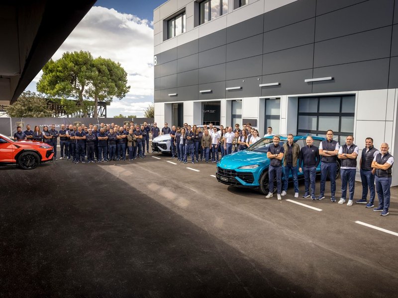 Large group of Lamborghini employees standing in front of the Sant'Agata Bolognese facility with three Lamborghini Urus models in orange, blue, and white