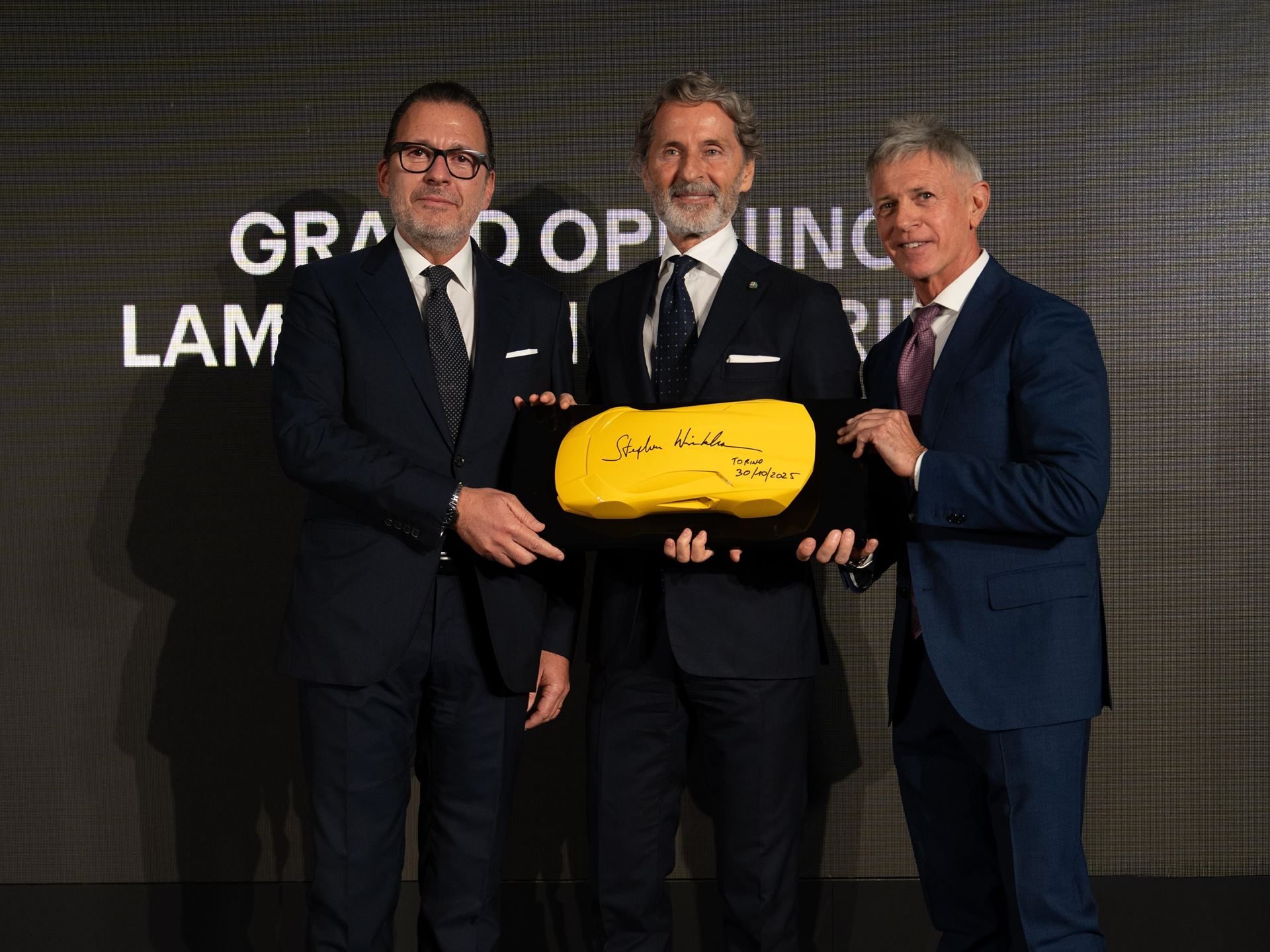 Lamborghini'S Seventh Italian Showroom Opens In Turin With 1,400 Square Meters And A 10,000-Rpm Centerpiece 3 Three men in suits holding a yellow car-shaped plaque at the lamborghini torino grand opening