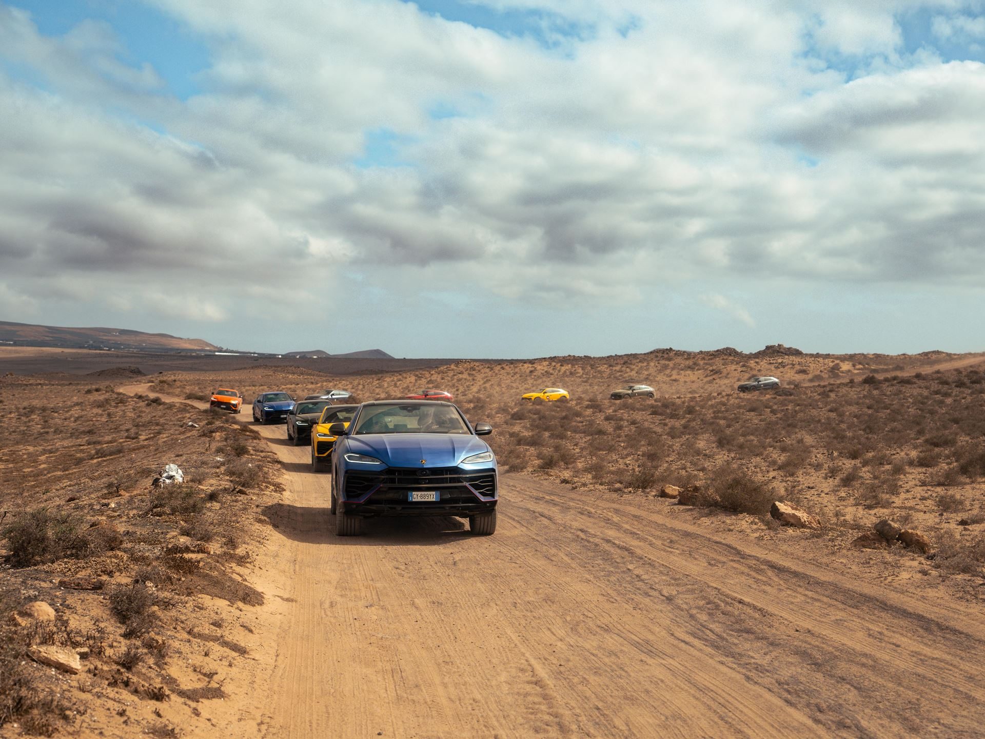Lanzarote'S Volcanic Terrain Becomes The Proving Ground For Lamborghini'S Plug-In Hybrid Urus Se 13 A convoy of blue, orange, black, and yellow lamborghini urus suvs driving on a dusty unpaved road through barren desert terrain