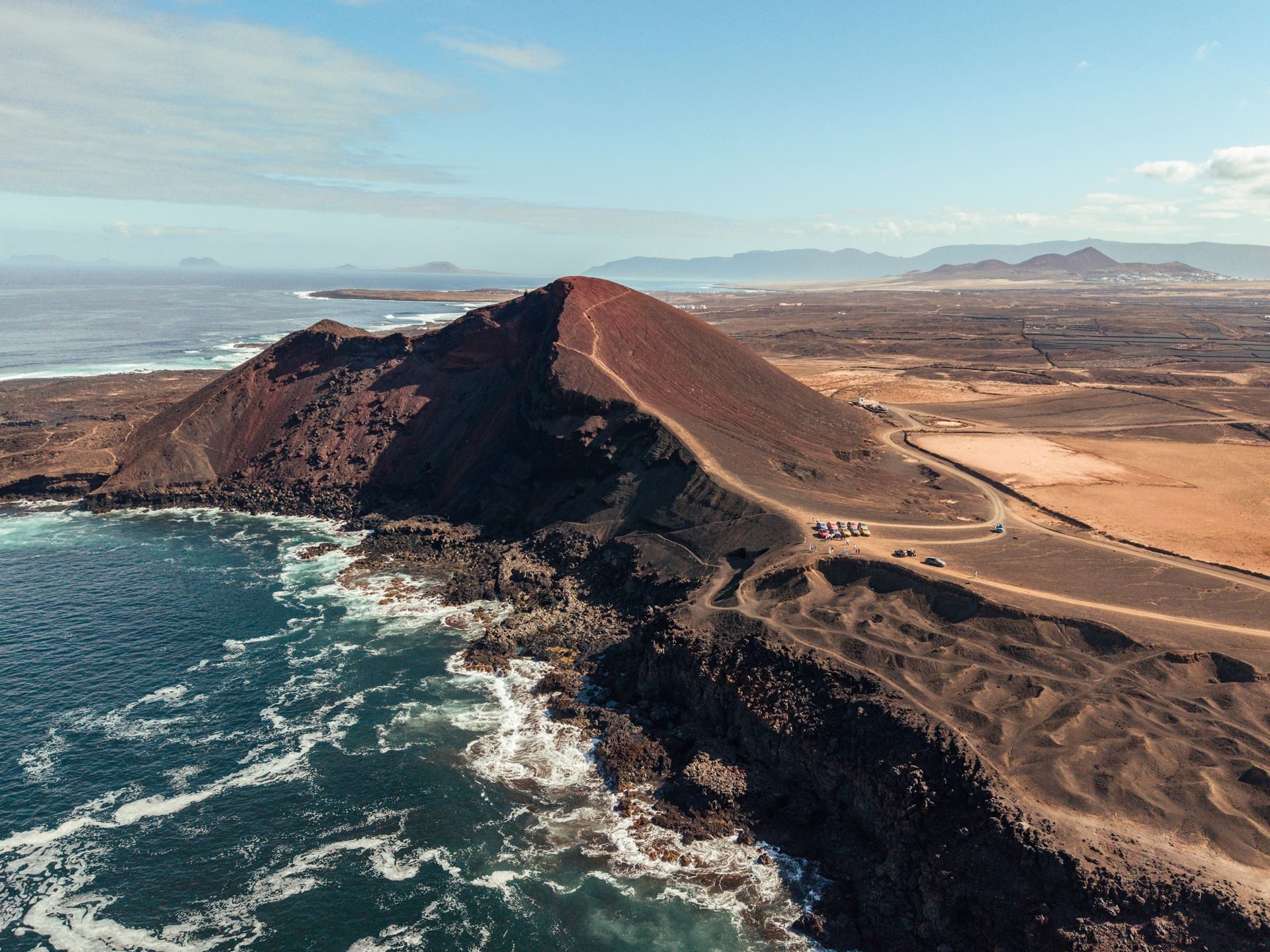 Lanzarote'S Volcanic Terrain Becomes The Proving Ground For Lamborghini'S Plug-In Hybrid Urus Se 9 Aerial view of lanzarote's volcanic coastline with a red-brown crater meeting the ocean and a dirt road leading to parked cars