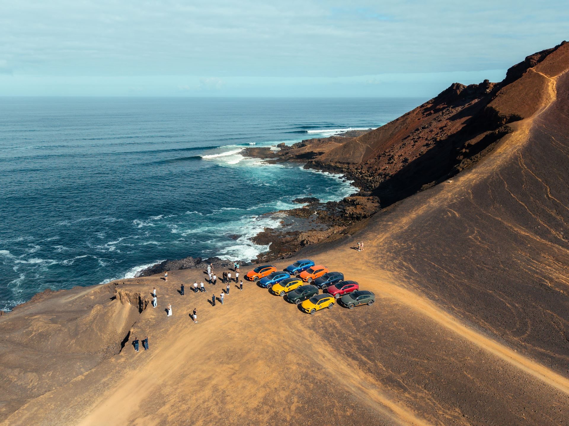 Lanzarote'S Volcanic Terrain Becomes The Proving Ground For Lamborghini'S Plug-In Hybrid Urus Se 11 Aerial view of colorful lamborghini urus suvs parked on a cliff edge overlooking the ocean with people gathered nearby