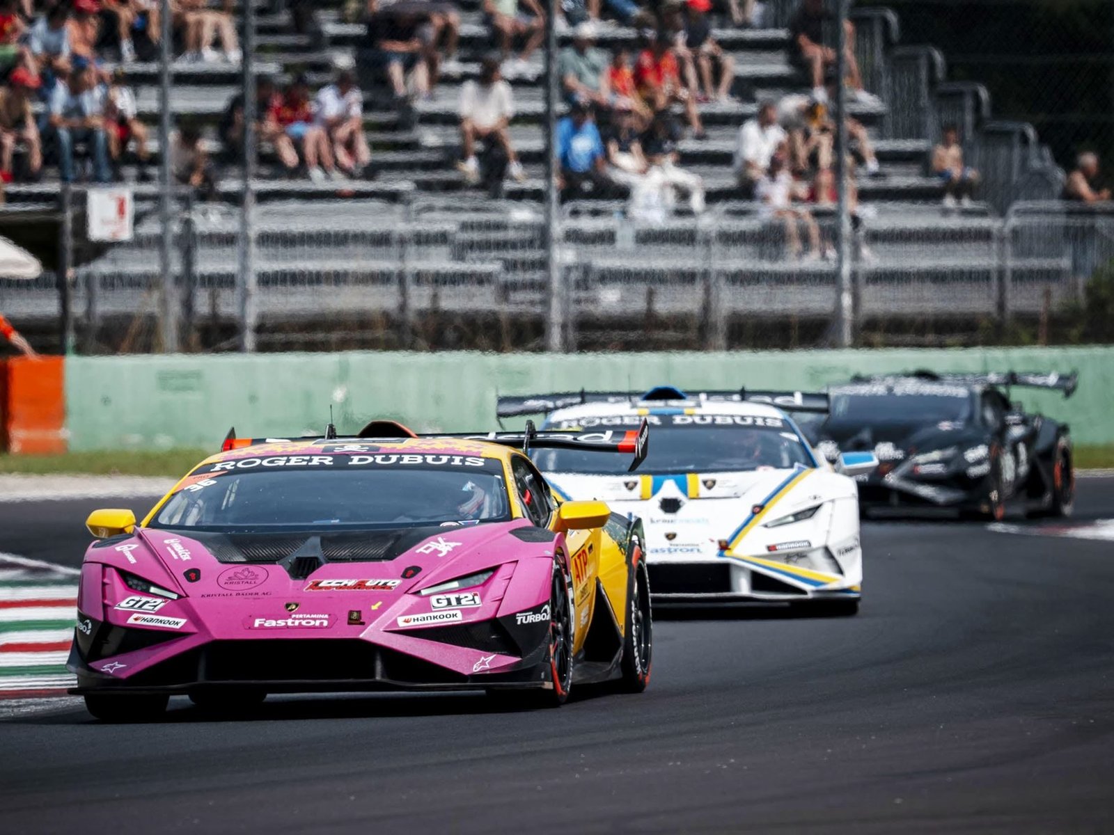 A pink and yellow Lamborghini Huracán Super Trofeo EVO2 leads two other race cars on track during a Super Trofeo race