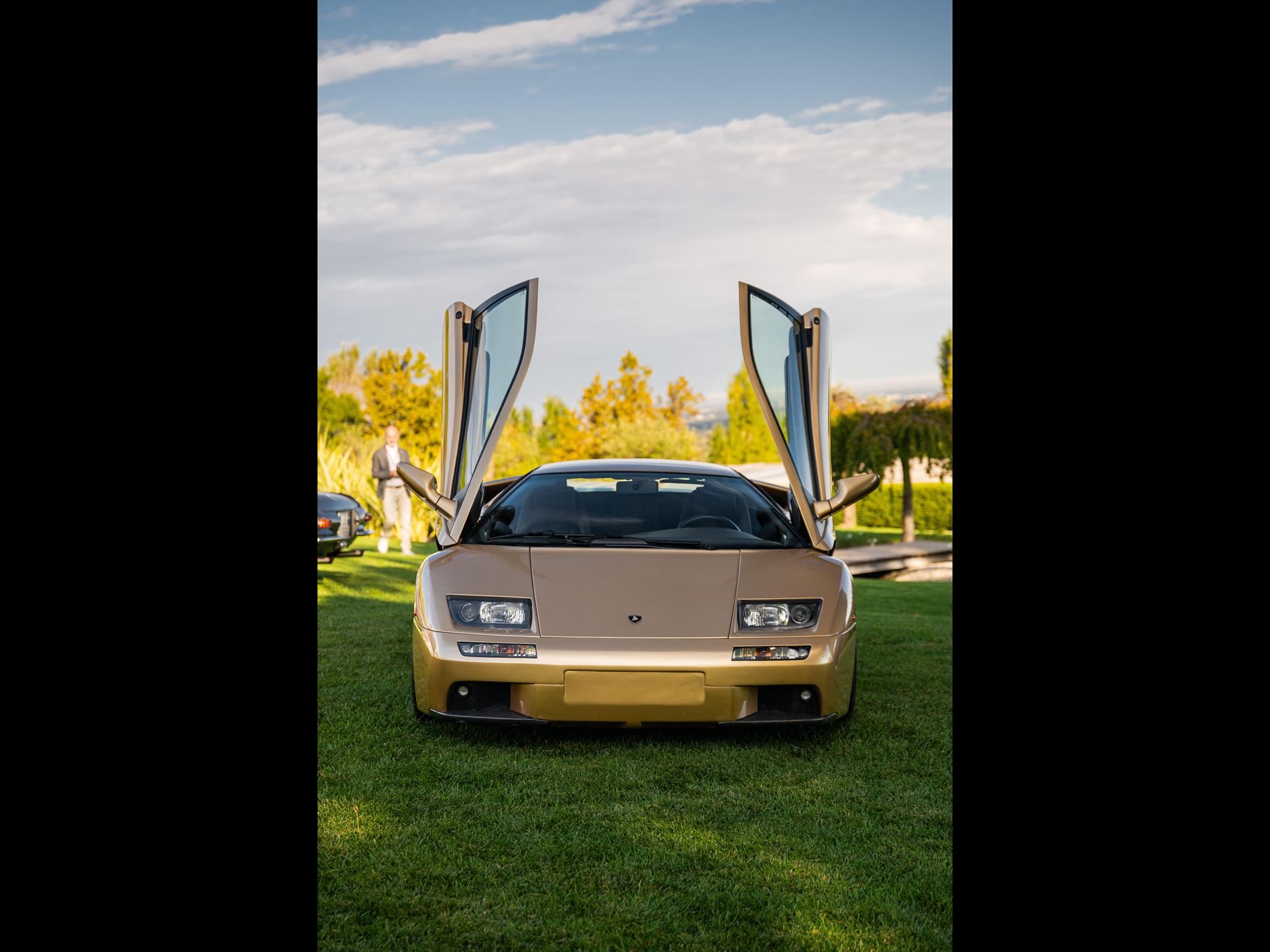 Gold lamborghini diablo vt with scissor doors open on green grass at an outdoor concours event under blue sky
