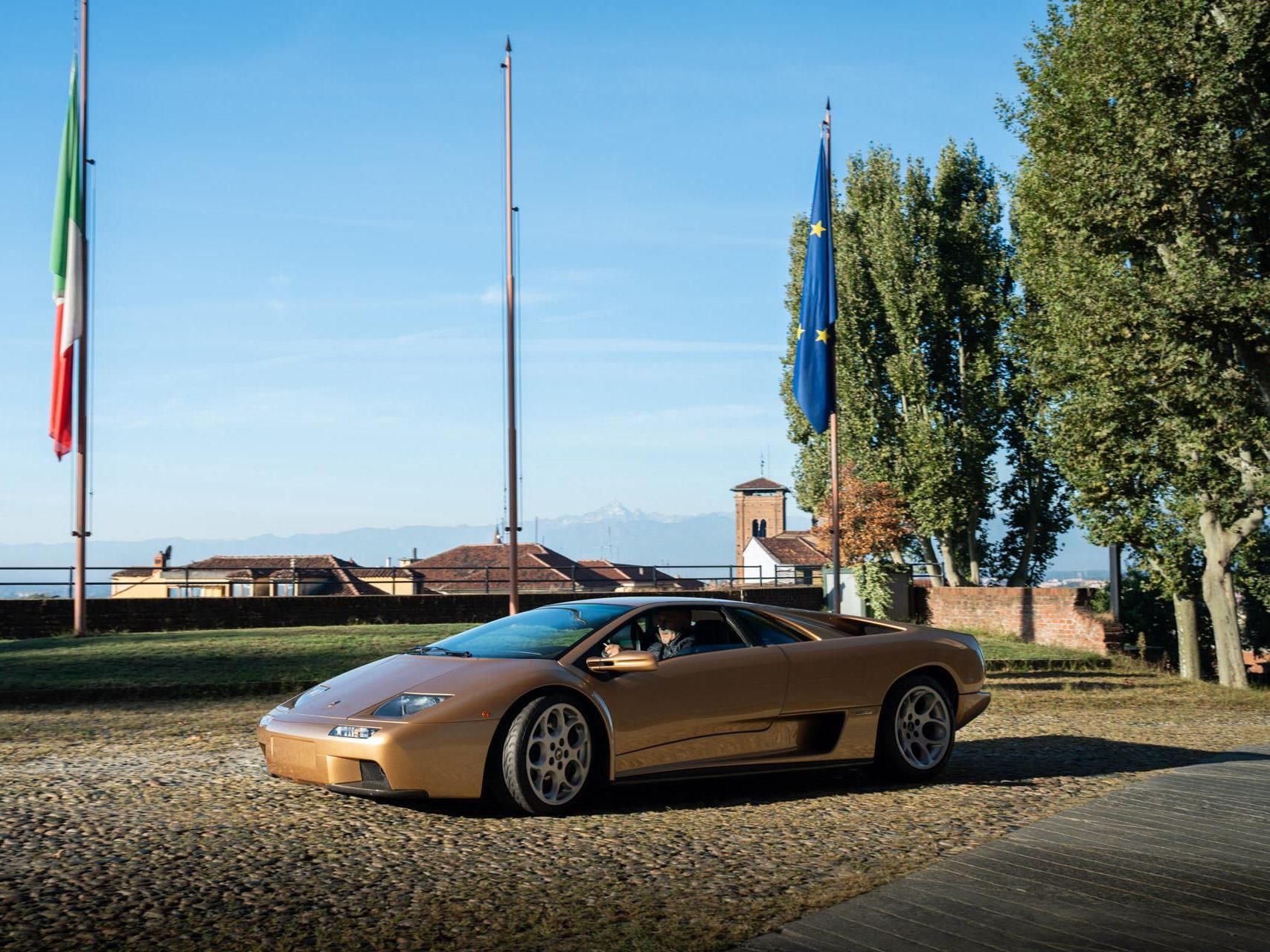 Gold lamborghini diablo 6. 0 vt driving on a cobblestone road with italian flags, trees, and distant mountains in the background