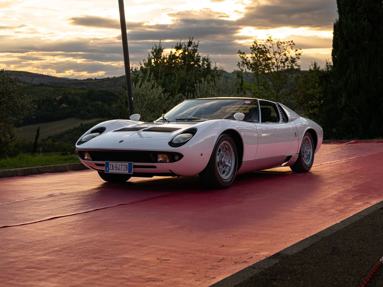 White Lamborghini Miura P400 parked on red carpet with dramatic sunset sky and rolling Italian hills in the background
