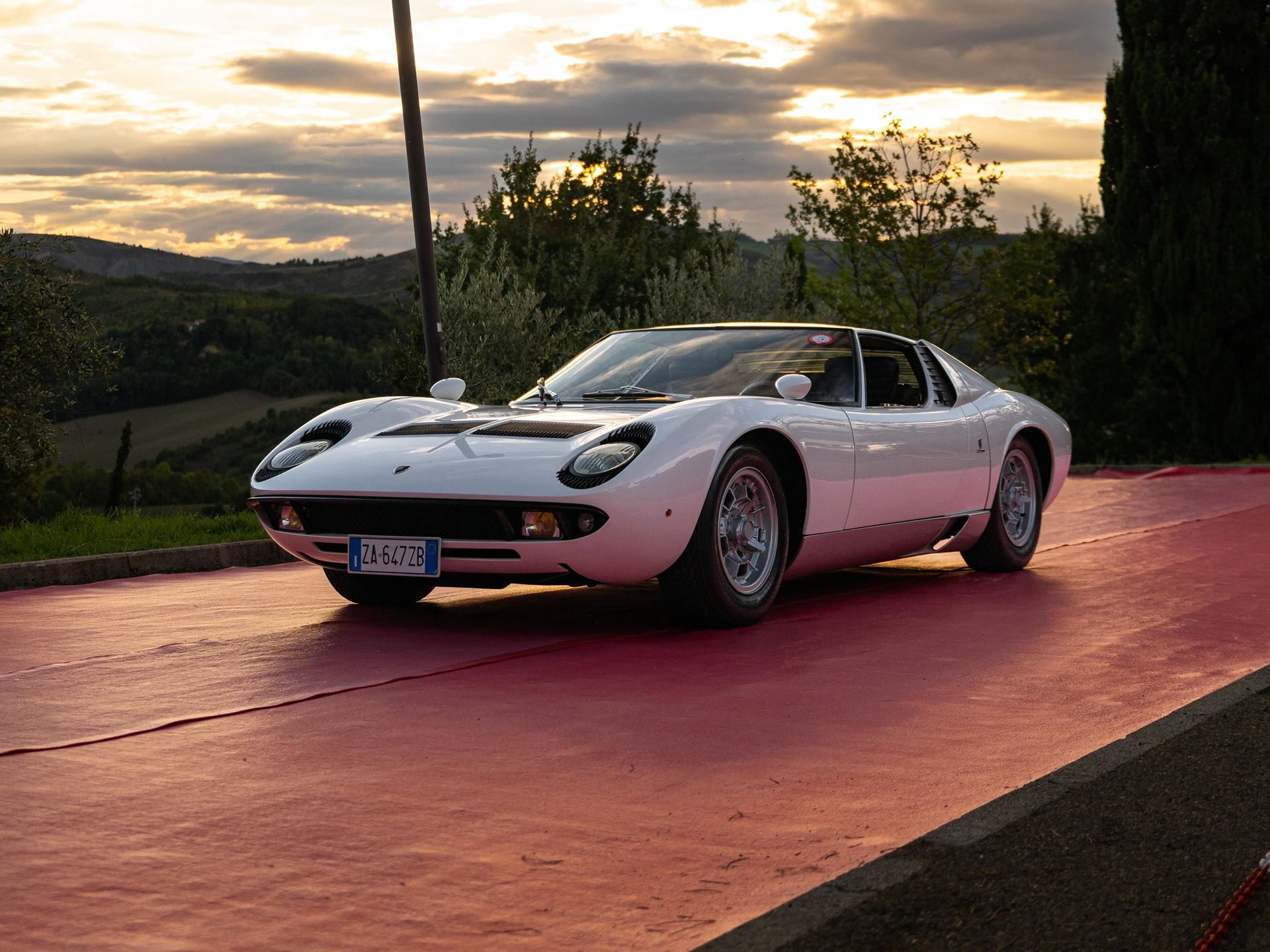 White lamborghini miura p400 parked on red carpet with dramatic sunset sky and rolling italian hills in the background