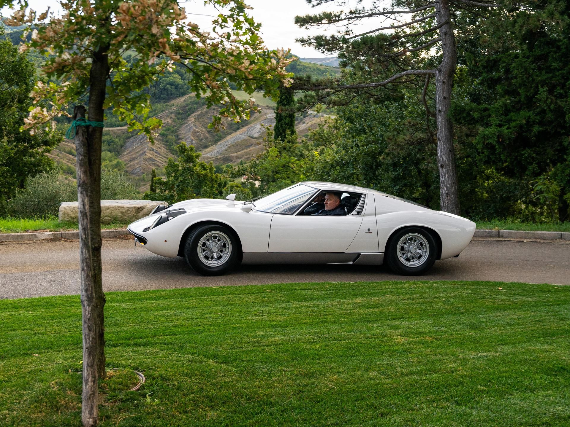 White lamborghini miura p400 driving on a winding road through scenic italian hills with trees and greenery
