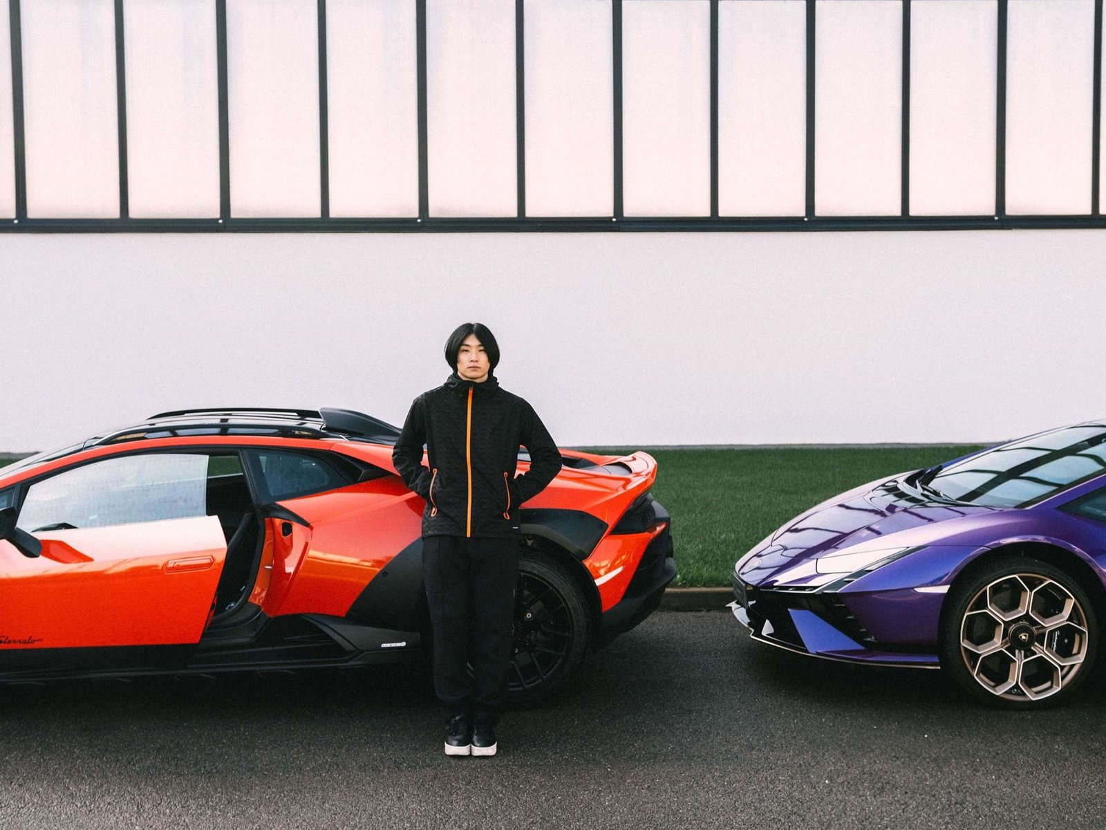 Model in black and orange Macron Lamborghini activewear standing between an orange Huracan Sterrato and a purple Huracan Tecnica