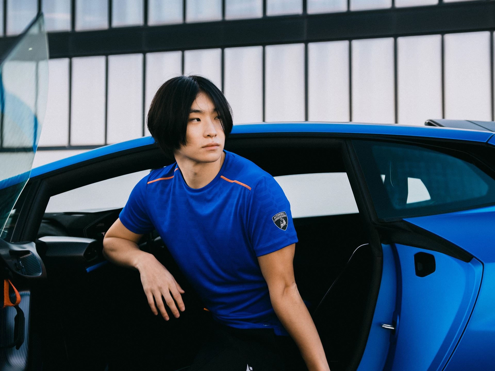Lamborghini'S Second Macron Activewear Drop Stitches Car-Seat Hexagons Into Running Gear 11 Model in blue macron lamborghini activewear with orange accents stepping out of a blue lamborghini huracan sto