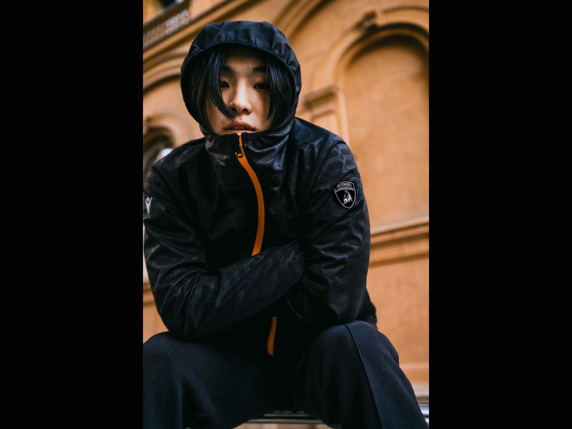 Lamborghini'S Second Macron Activewear Drop Stitches Car-Seat Hexagons Into Running Gear 9 Person wearing black lamborghini macron activewear jacket with orange accents and lamborghini shield logo, seated against a brick background