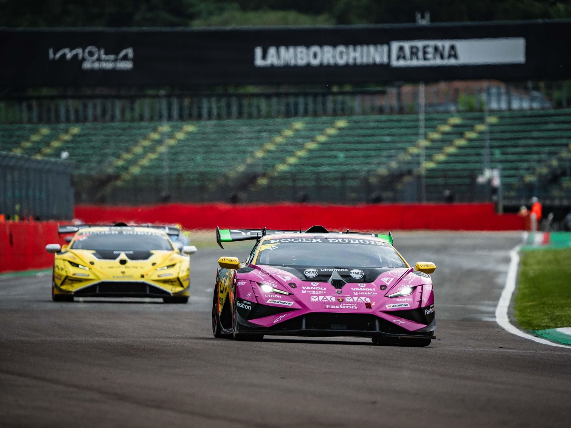 Oregon team's pink and yellow #36 huracán super trofeo evo2 leads a yellow competitor down the straight under the lamborghini arena banner at imola