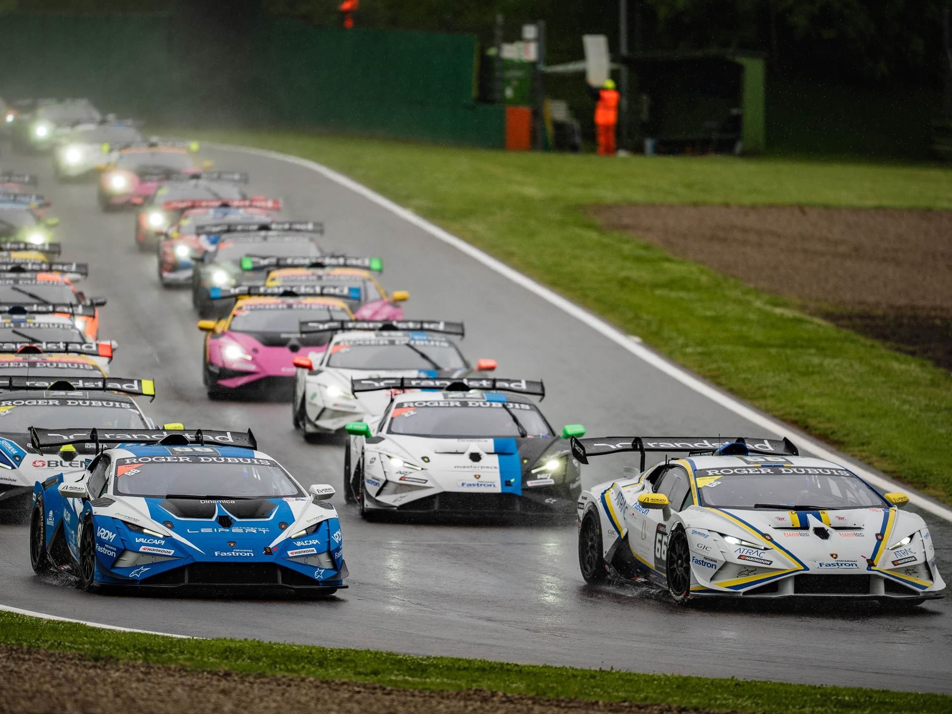 Multiple huracán super trofeo evo2 cars race through spray on a wet imola circuit, with two cars leading the pack in the foreground