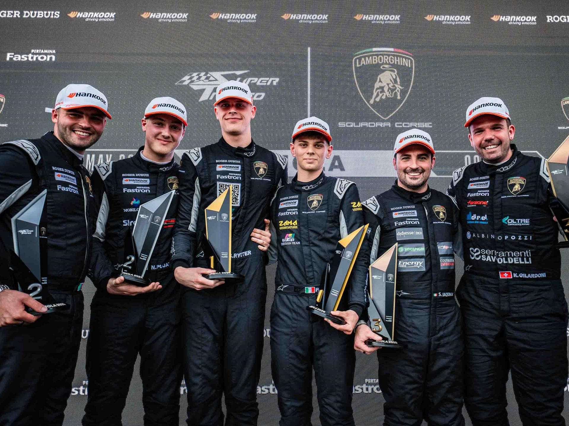 Six race car drivers hold trophies on the podium with the lamborghini squadra corse logo displayed on a large screen behind them