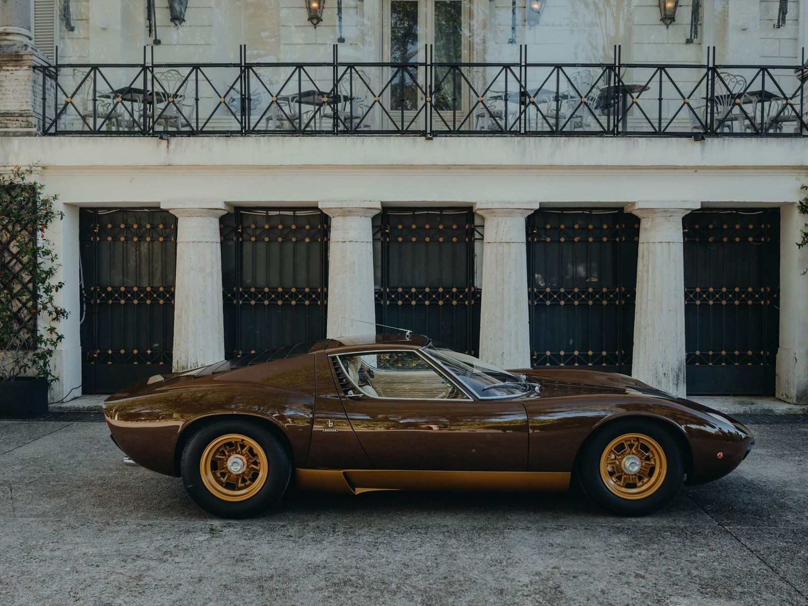 Side profile of the restored 1972 Lamborghini Miura SV in Luci del Bosco brown with gold wheels, parked in front of a classical columned building