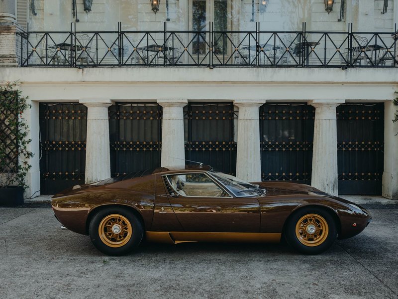 Side profile of the restored 1972 Lamborghini Miura SV in Luci del Bosco brown with gold wheels, parked in front of a classical columned building