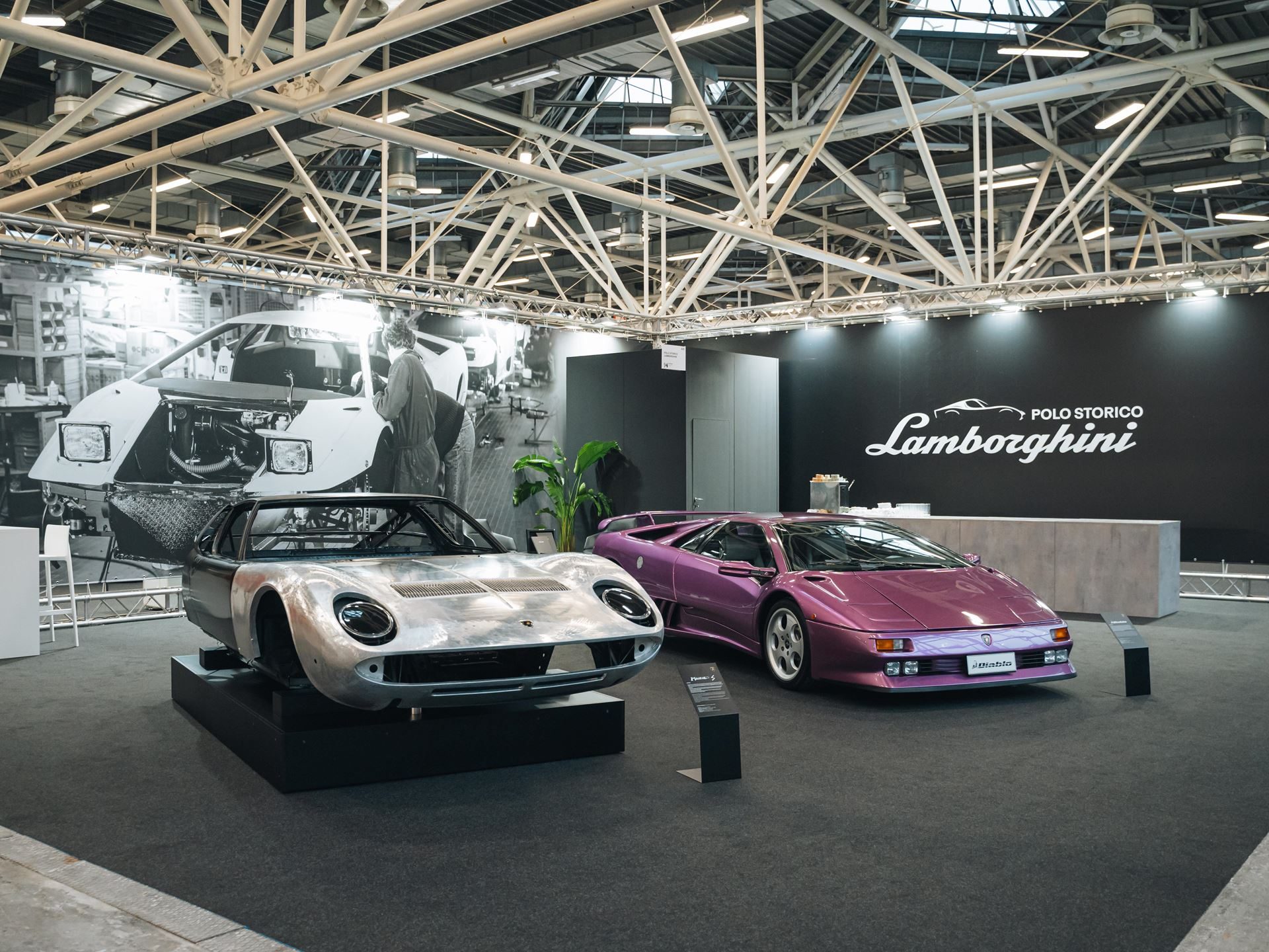 A wide shot of the Lamborghini Polo Storico stand at an automotive event, featuring a silver Miura S chassis and a purple Diablo SE30, with a large historical backdrop and event branding.