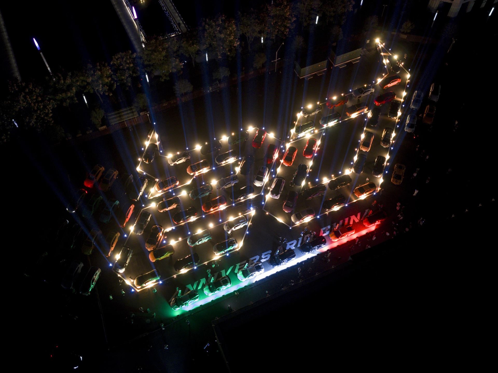 Aerial night view of 100 lamborghini cars arranged to spell '20 years riding in china' with italian flag colors against shanghai skyline