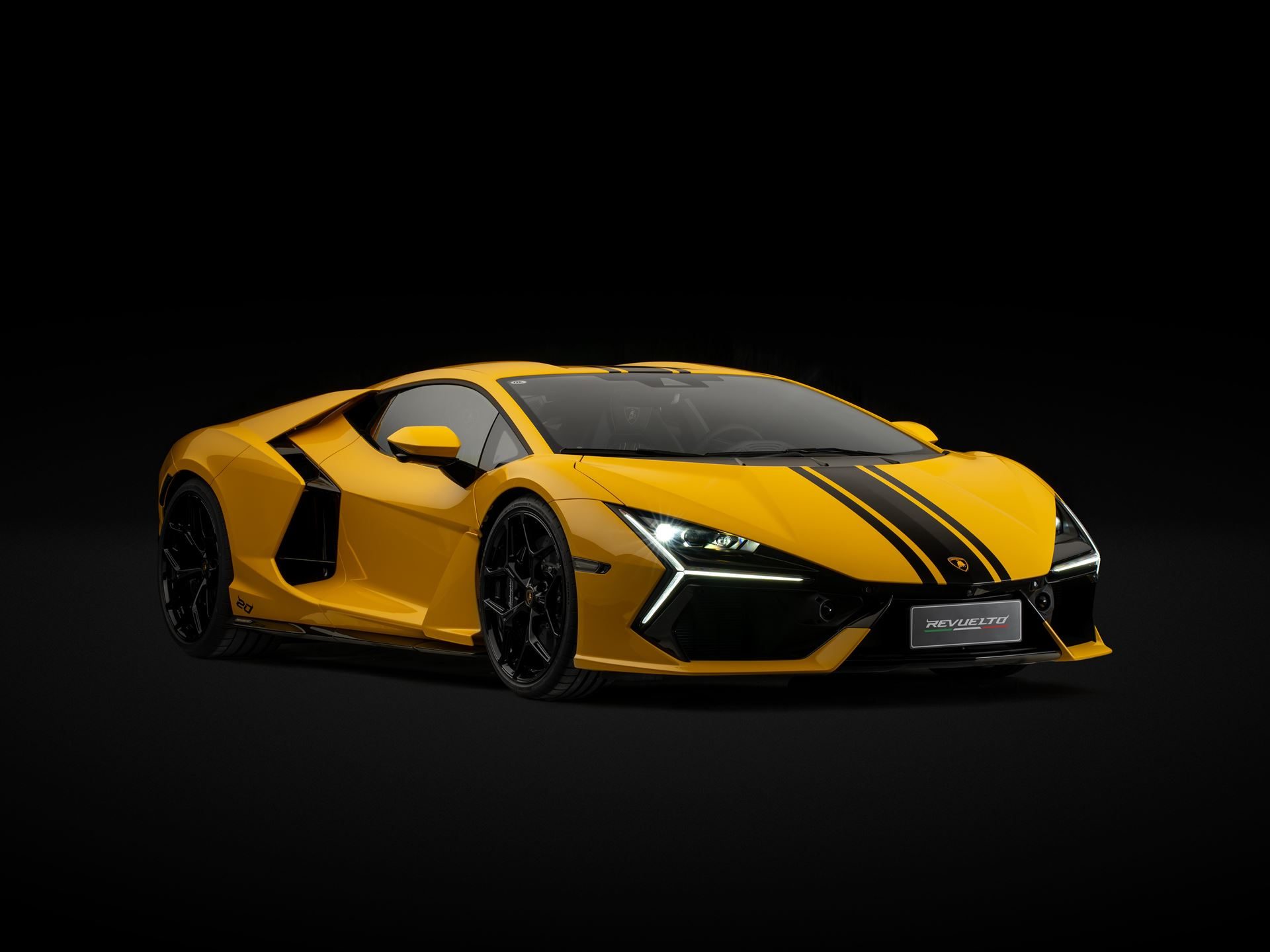 Front three-quarter view of the yellow lamborghini revuelto ad personam 20th anniversary with black racing stripes and illuminated headlights