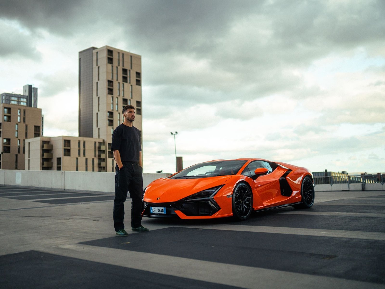 Rúben Dias standing beside an orange Lamborghini Revuelto on an urban rooftop