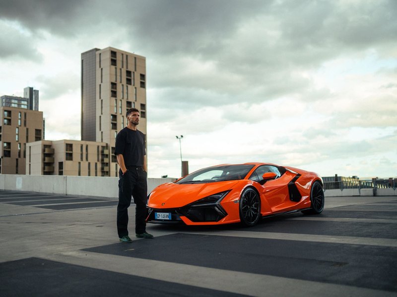 Rúben Dias standing beside an orange Lamborghini Revuelto on an urban rooftop
