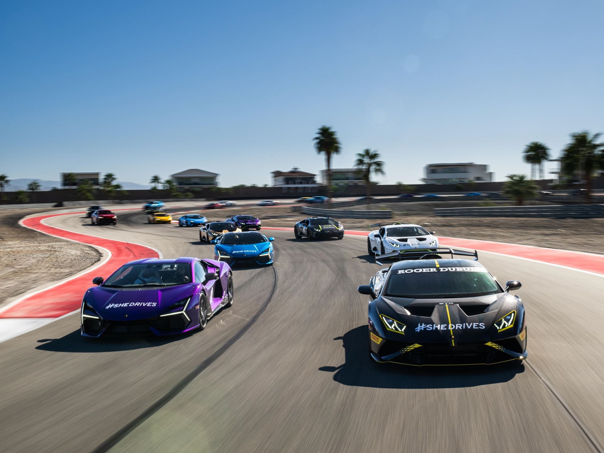 Shedrives A Lambo Is Not Just A Feel-Good Event. It Is Lamborghini'S Play For Long-Term Loyalty. 7 Group of lamborghini supercars including a purple revuelto and black huracán sto in motion on a racetrack with palm trees in the background