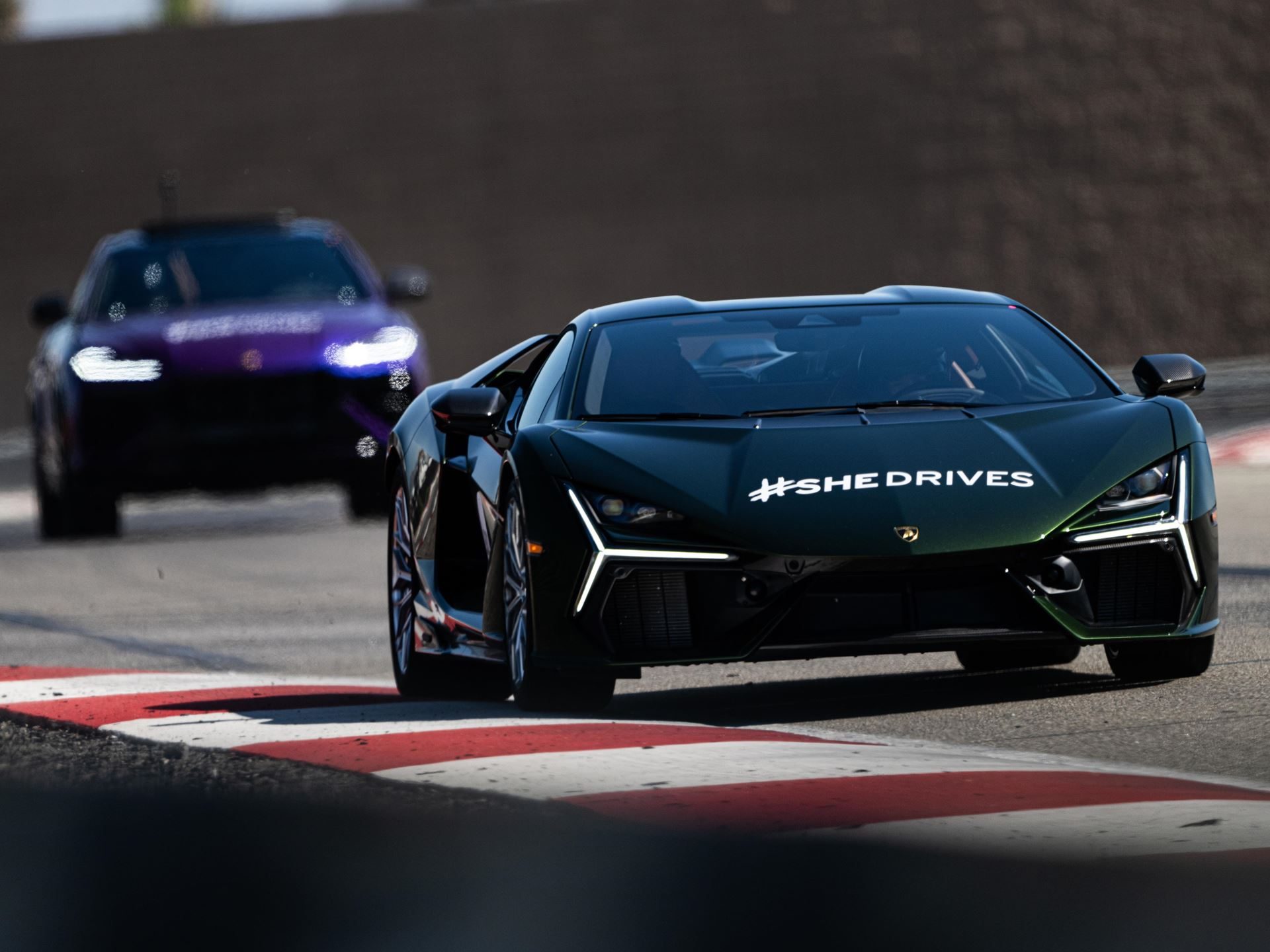 Shedrives A Lambo Is Not Just A Feel-Good Event. It Is Lamborghini'S Play For Long-Term Loyalty. 9 Dark green lamborghini revuelto with #shedrives branding leading on a racetrack with a purple urus following behind