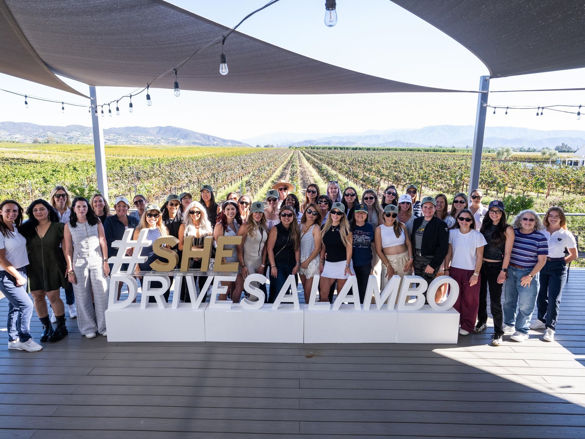 Shedrives A Lambo Is Not Just A Feel-Good Event. It Is Lamborghini'S Play For Long-Term Loyalty. 3 Large group of women posing in front of a #shedrivesalambo sign on a wooden deck with a vineyard in the background
