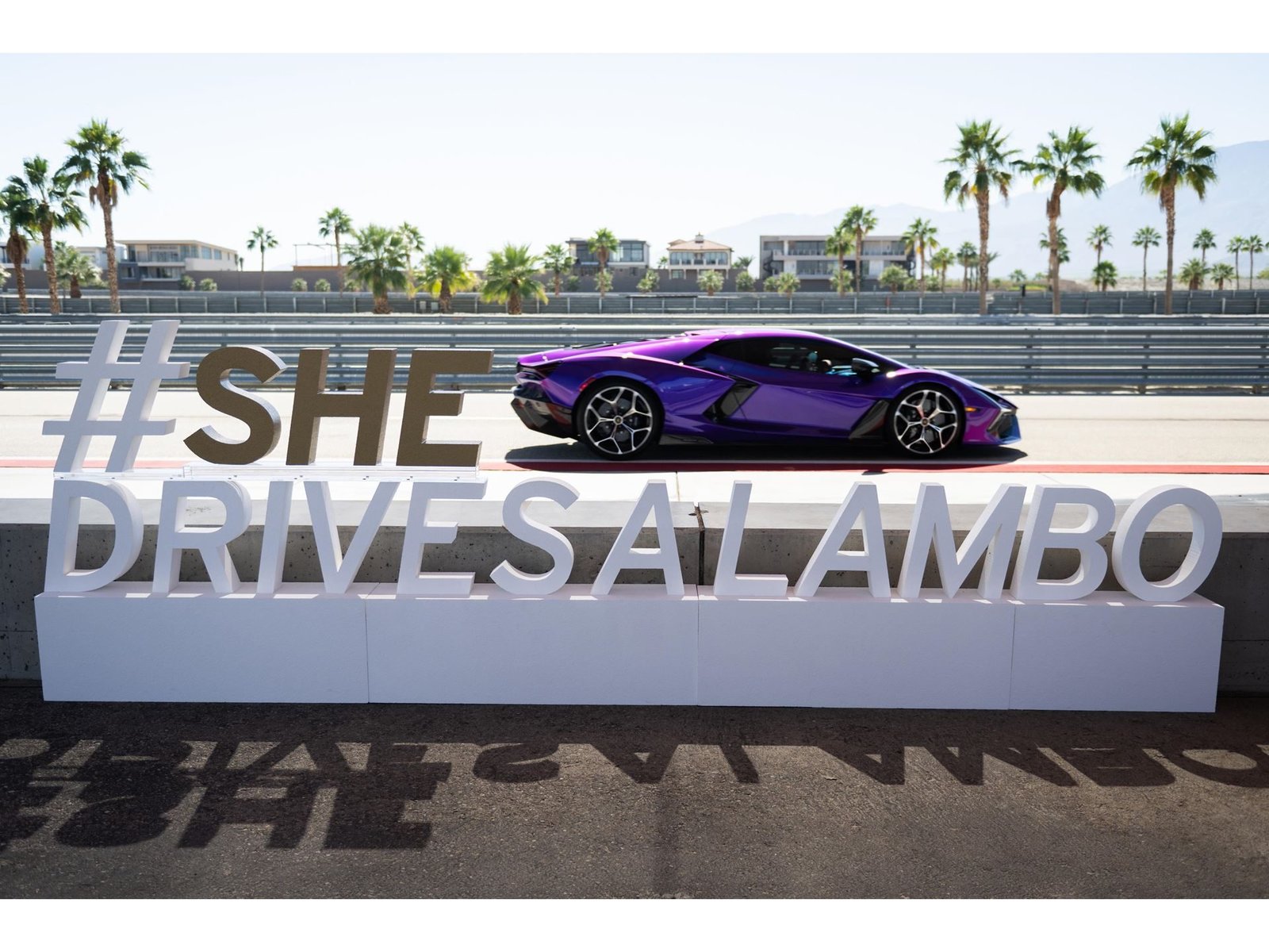 Purple Lamborghini Revuelto parked on the racetrack at The Thermal Club next to a large #SHEDRIVESALAMBO sign with palm trees in the background