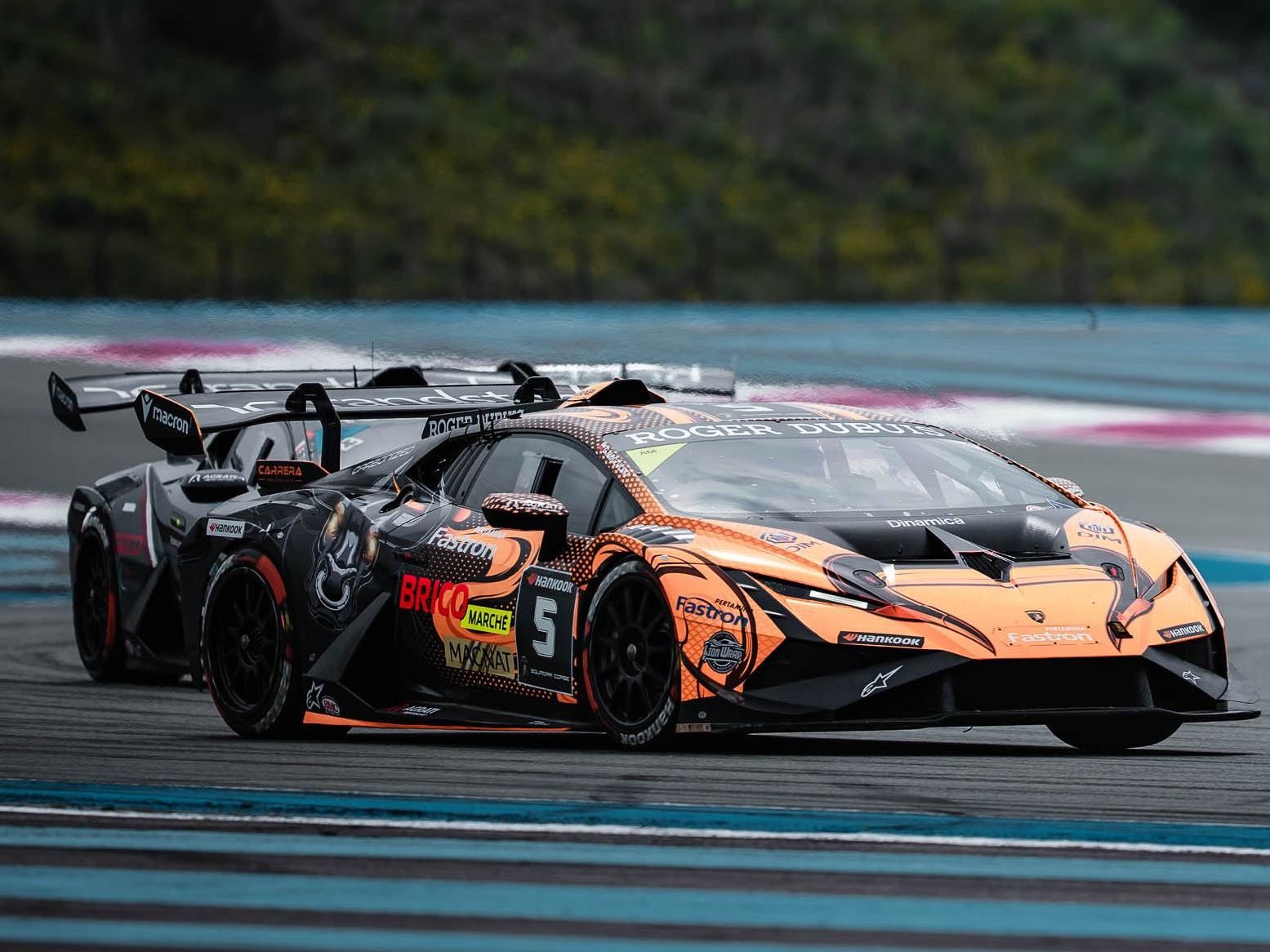 An orange and black liveried lamborghini huracán super trofeo evo2 number 5 leading another race car with prominent front splitter and rear wing visible