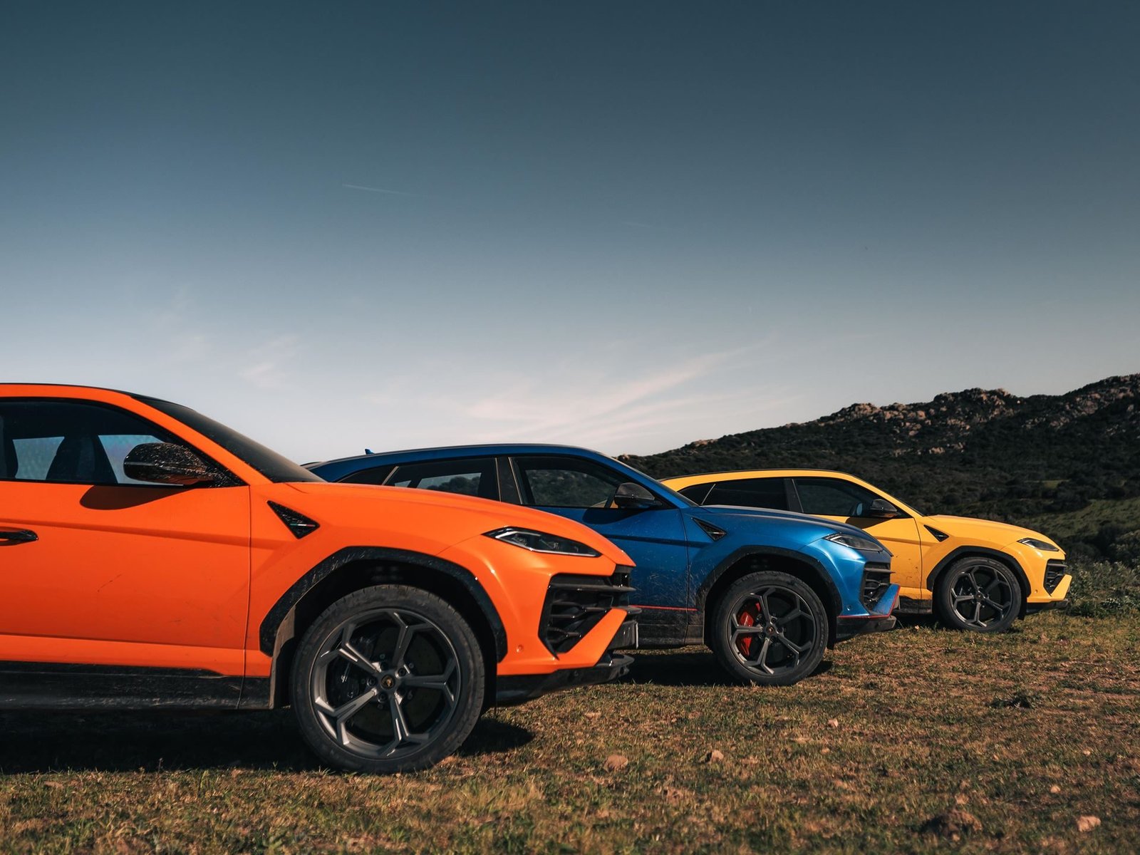 Three Lamborghini Urus SUVs in orange, blue, and yellow parked side-by-side on a grassy Sardinian hilltop