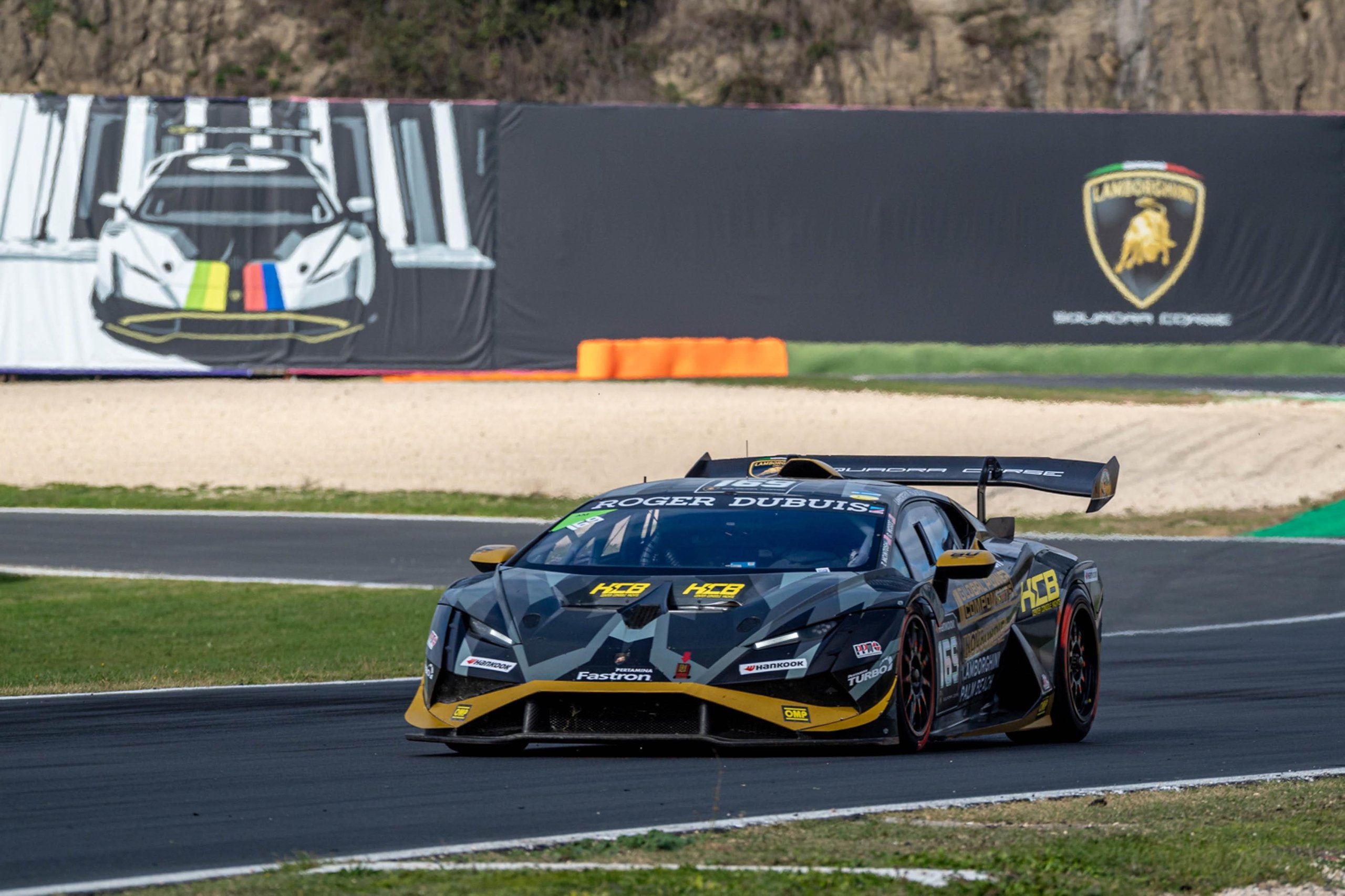 Black and gold lamborghini huracán super trofeo evo2 on a straight at vallelunga with a large lamborghini logo visible in the background